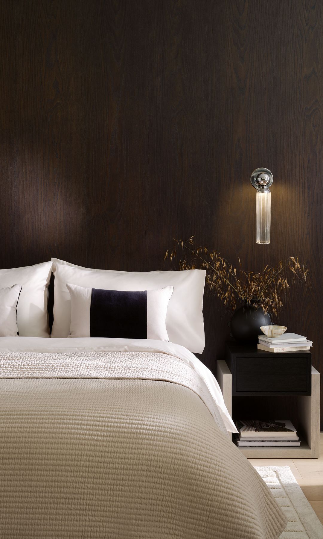 Dormitorio con pared del cabecero de madera, sábanas blancas, cojines negros, mesita de noche, aplique de pared sobre la mesita de noche