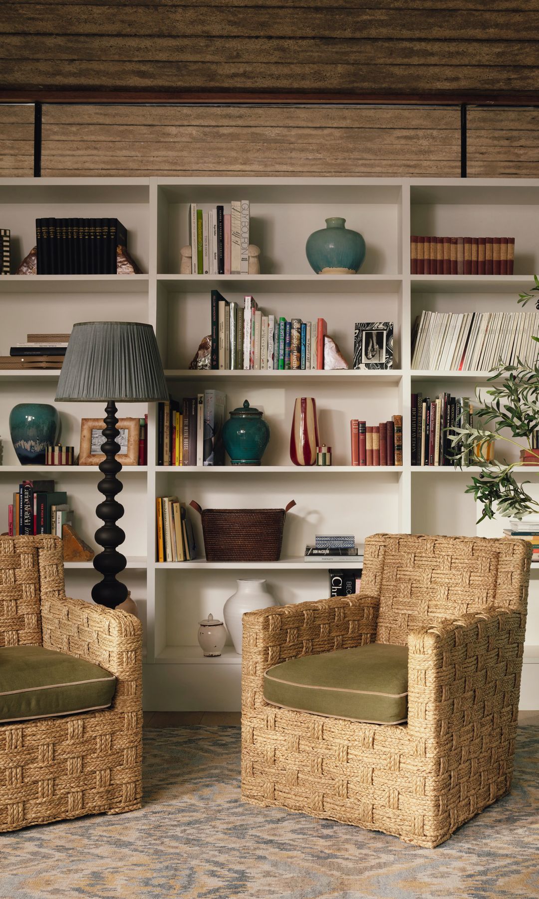 Salón con estantería blanca en la pared, libros y adornos, sillones de fibras vegetales, lámpara de pie, macetas, 