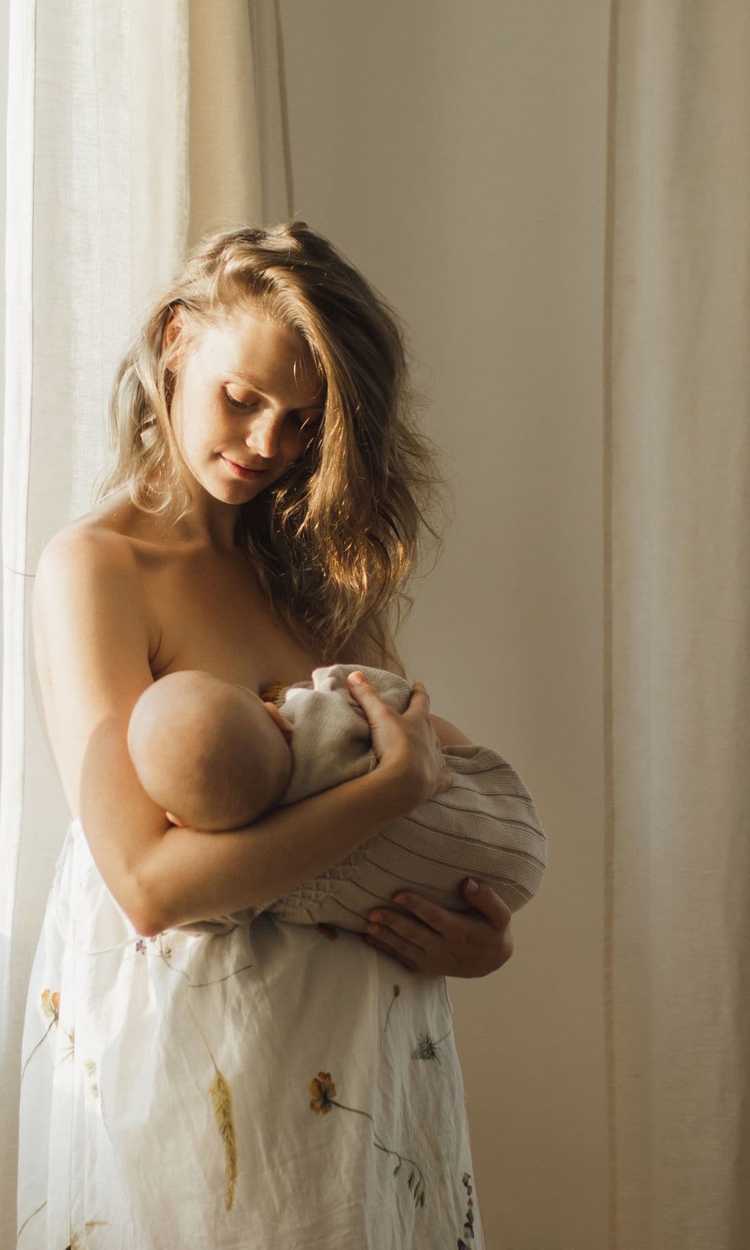 Mujer da el pecho a su bebé junto a la ventana