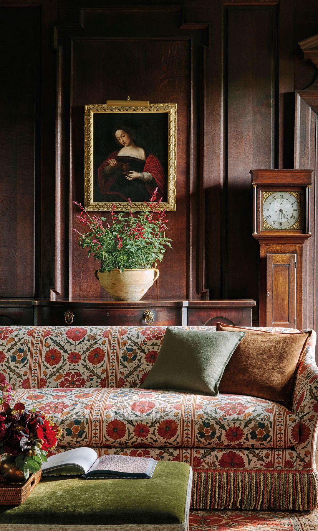 Salón con sofá tapizado con tela estampada de color rojo, paredes revestidas de madera oscura, reloj de pie, cuadro en la pared con marco dorado, arreglo floral, aparador de madera oscura, mesa de centro tapizada 