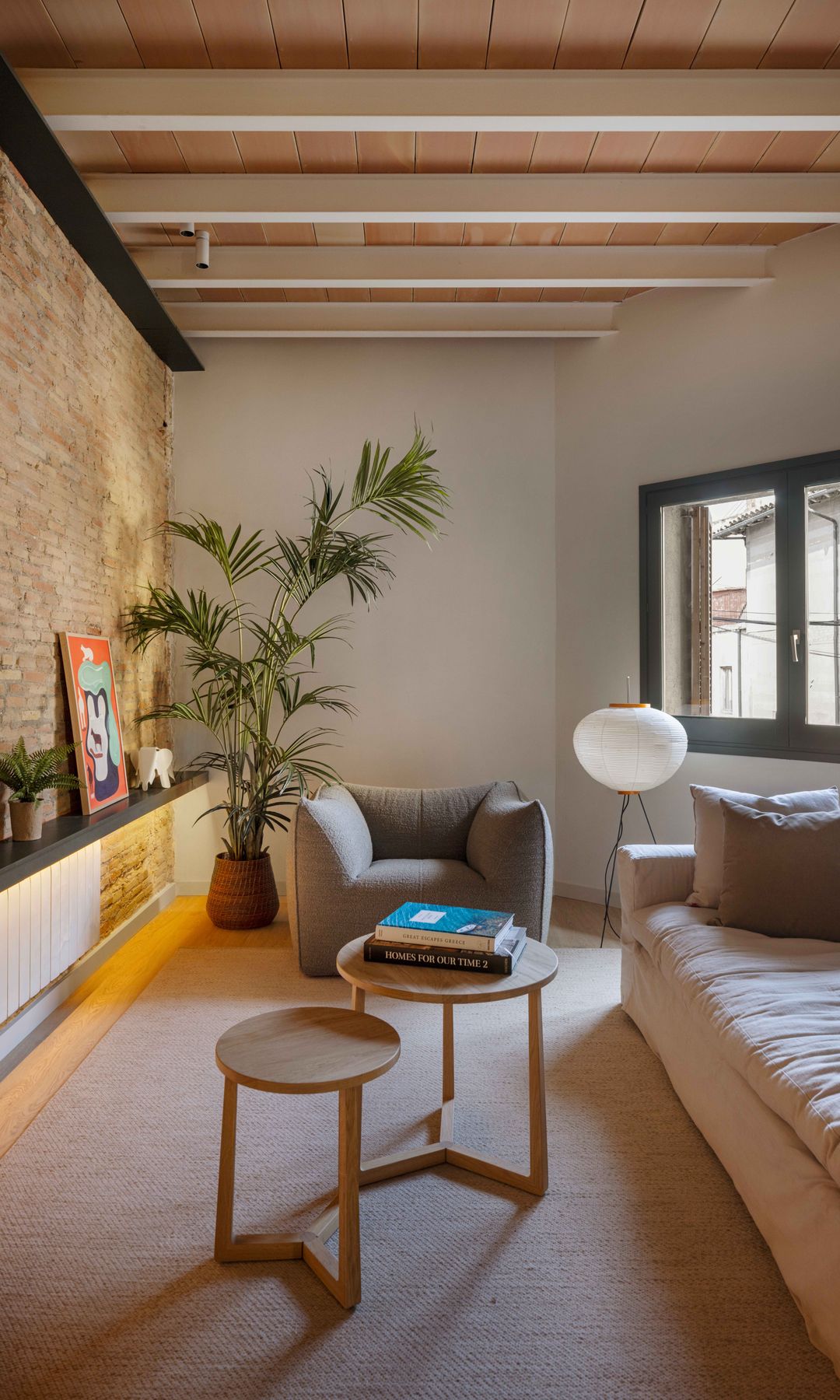 Salón de planta rectangular con pared de ladrillo, vigas de madera en el techo, sofá beige, mesas auxiliares de madera, planta XL en una maceta