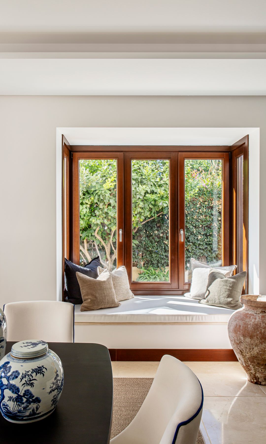 Rincón con banco bajo la ventana con almohadones blancos, cojines y ventanas con marco de madera