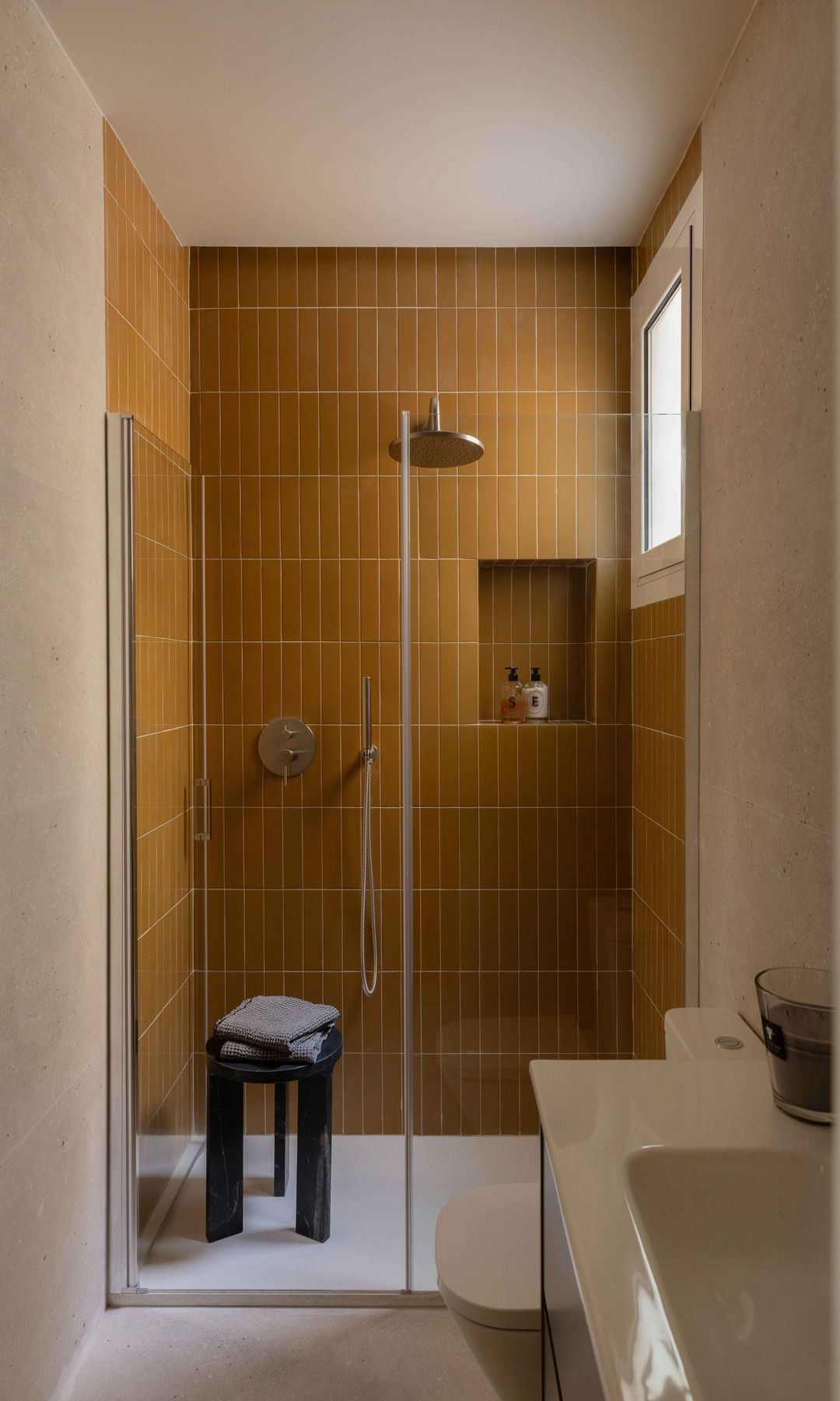 Baño con ventana, ducha revestida de azulejos marrones, plato de ducha blanco, mampara, lavabo blanco, taburete
