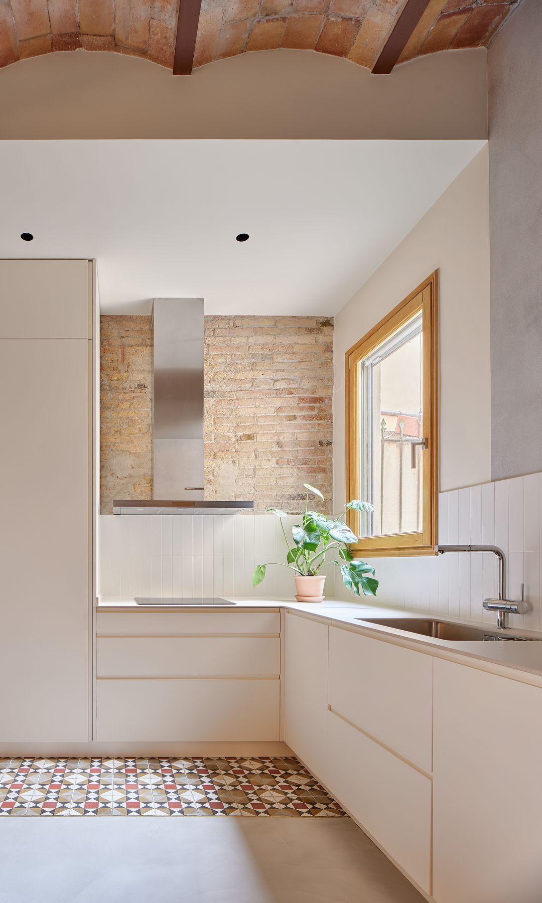Cocina blanca con suelo hidráulico, bóvedas catalanas en el techo, ventana de madera, pared de ladrillo visto, mesa de office de madera, silla con el respaldo de rejilla