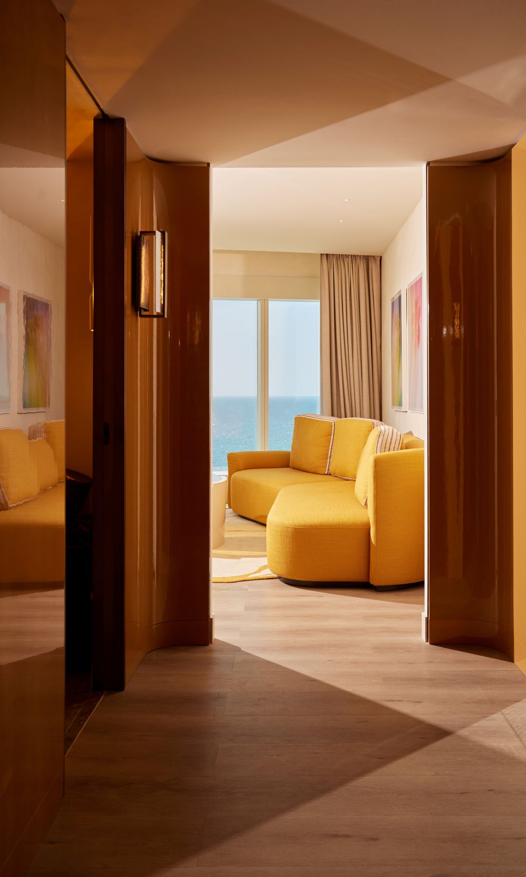 Habitación de hotel con gran ventanal, sofá amarillo, suelo de madera, espejo, apliques pared