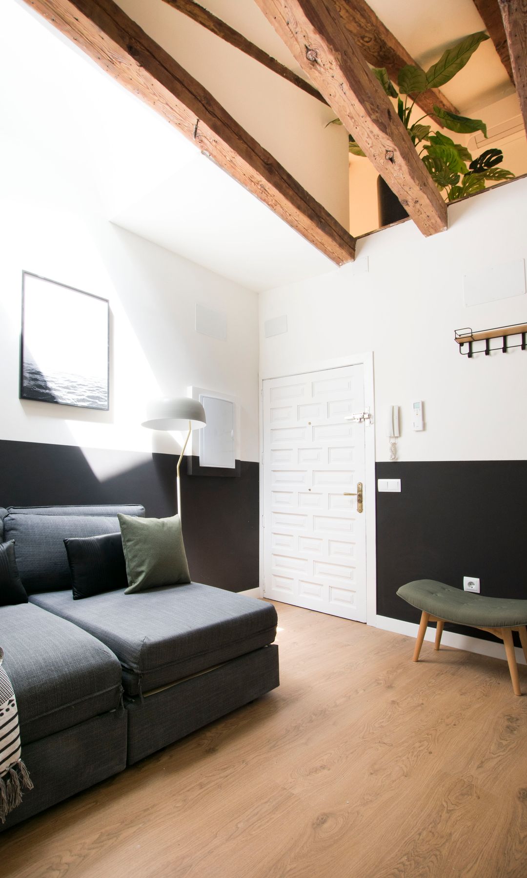 Salón pequeño con zócalo alto negro, sofá gris, entrada directa al salón con puerta blanca, vigas en el techo, suelo de madera, cuadros en la pared, taburete, perchero pared