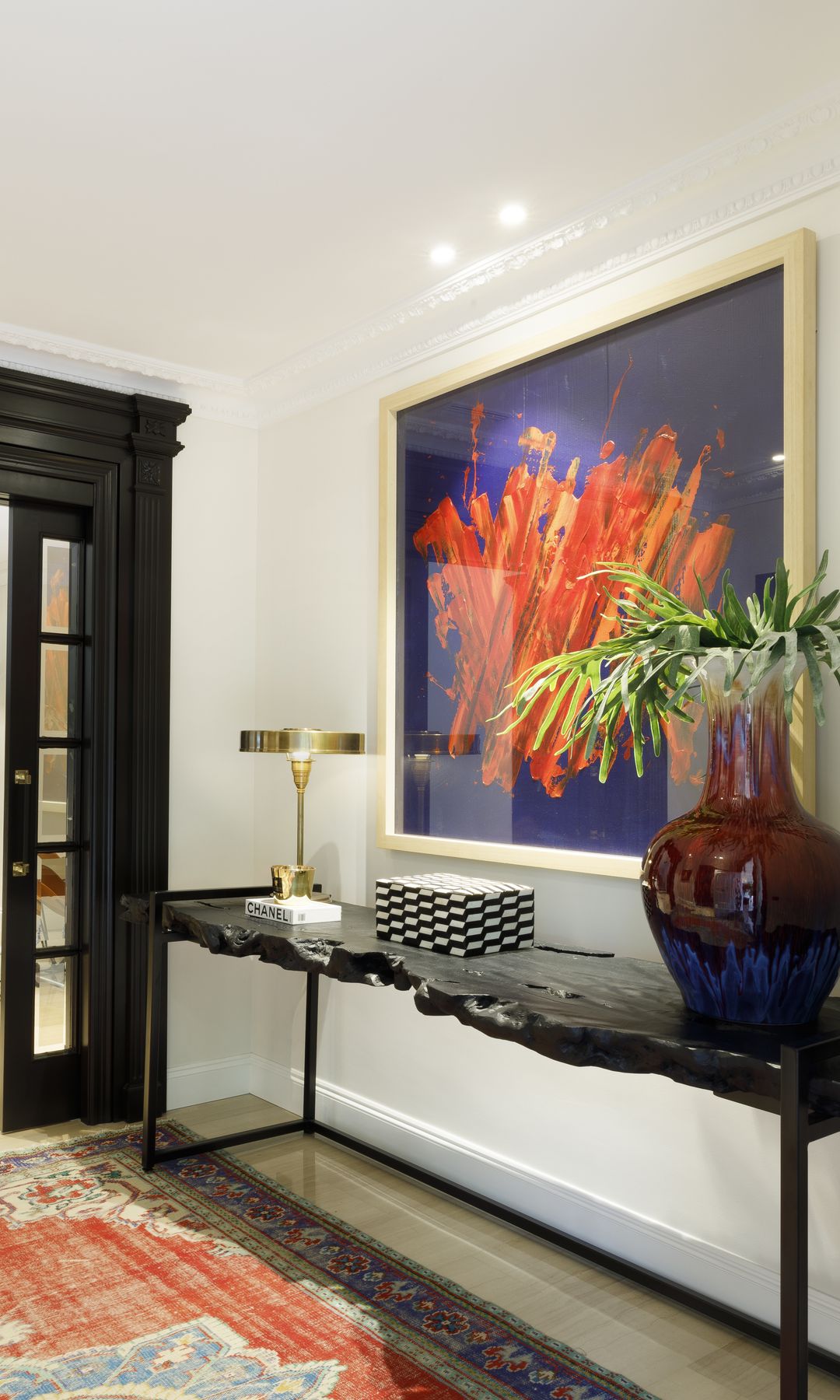 Recibidor con consola negra de mármol, cuadro en la pared, aplique, jarrón de cristal de colores, lámpara de sobremesa dorada, alfombra