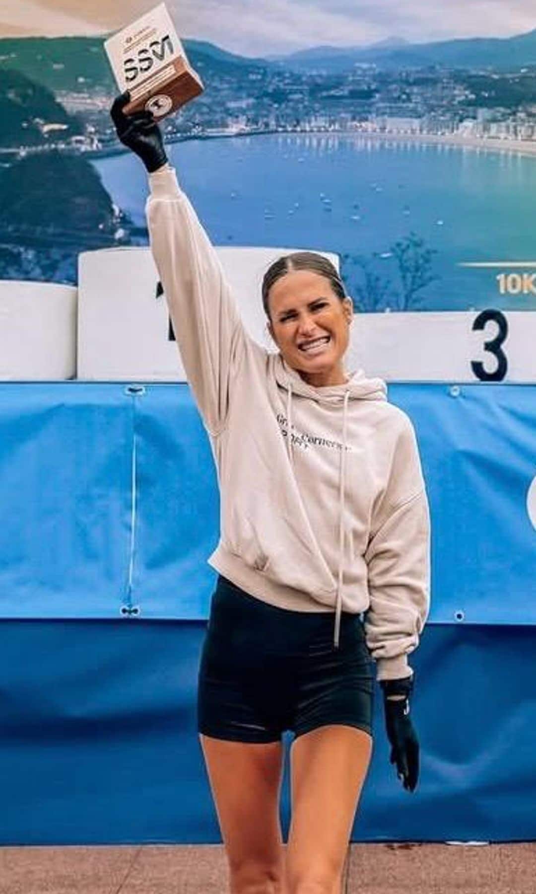 Verdeliss en hoodie y shorts, levantando una medalla con su mano derecha, sonriendo.