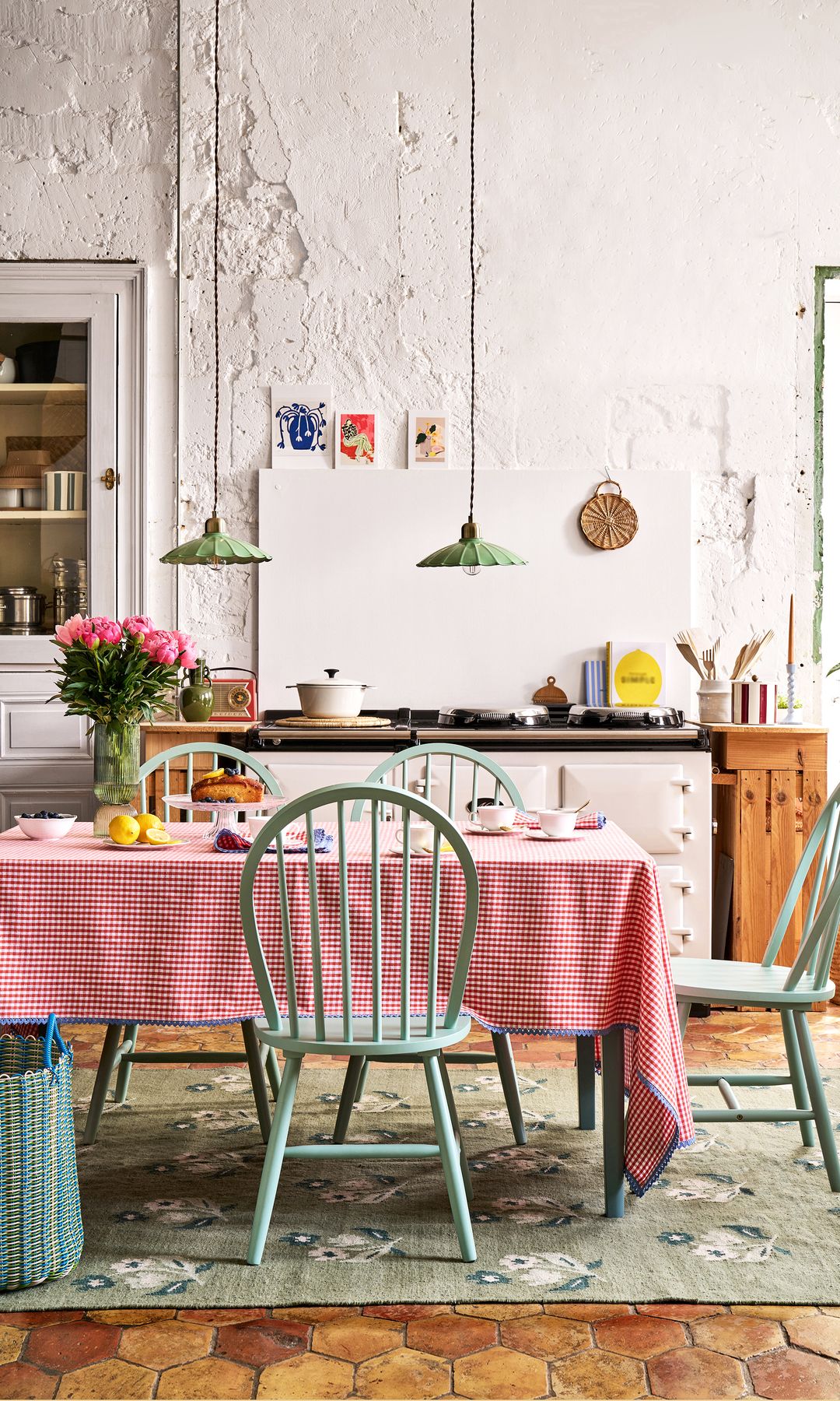 Cocina con encanto con mesa de comedor con mantel de cuadros rojos, alfombra, paredes encaladas, sillas verdes, plantas, jarrón con flores, lámparas de techo sobre la mesa 