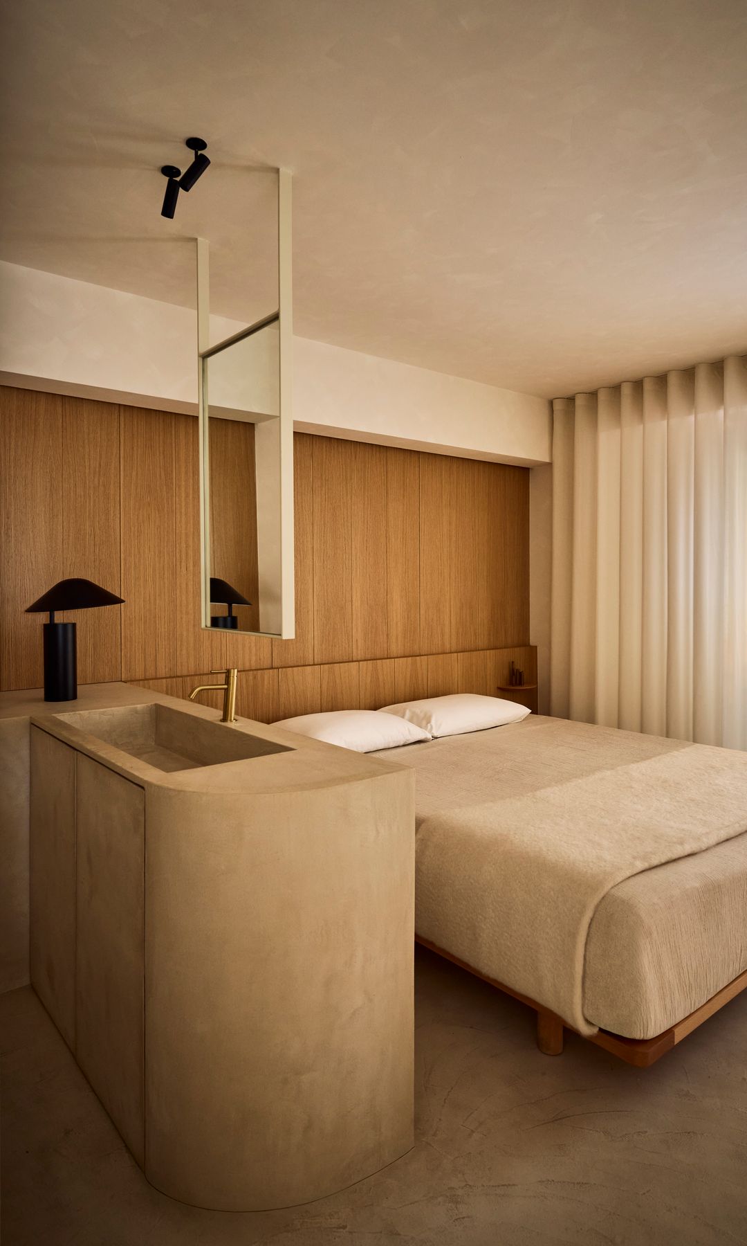 Dormitorio con baño integrado en la habitación con mueble de lavabo de microcemento, cabecero de madera de pared a pared, espejo de baño colgante