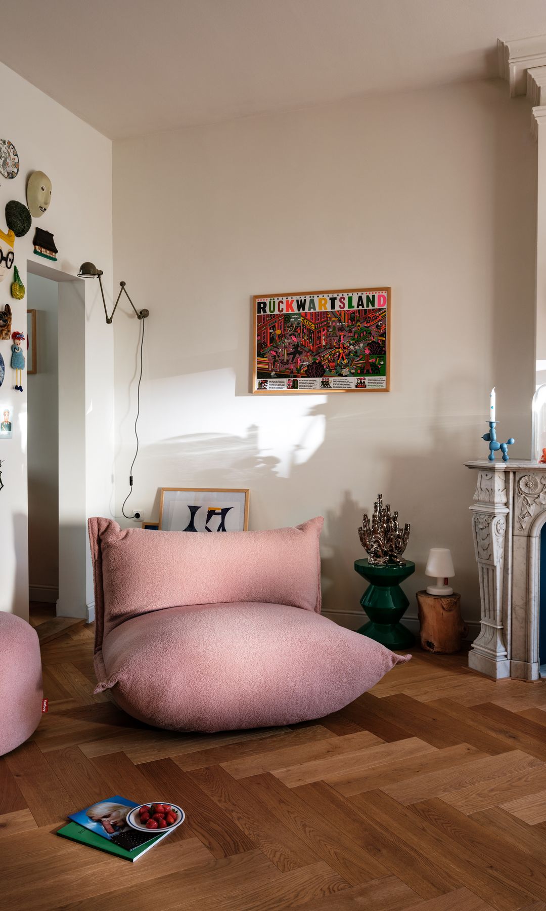 Salón con chimenea, suelo de madera de espiga, puff rosa con respaldo y puff redondo a juego, adornos en la pared, candelabros de colores sobre la chimenea