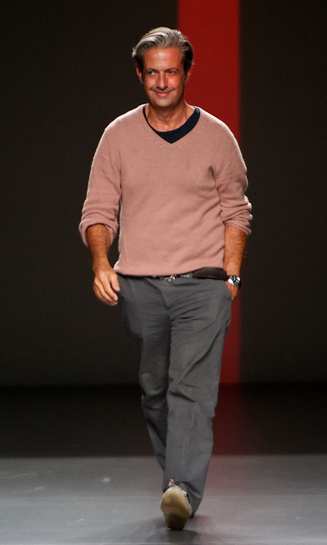 Miguel Palacio con cabello gris, sonriendo, caminando en una pasarela con traje gris y suéter rosa.