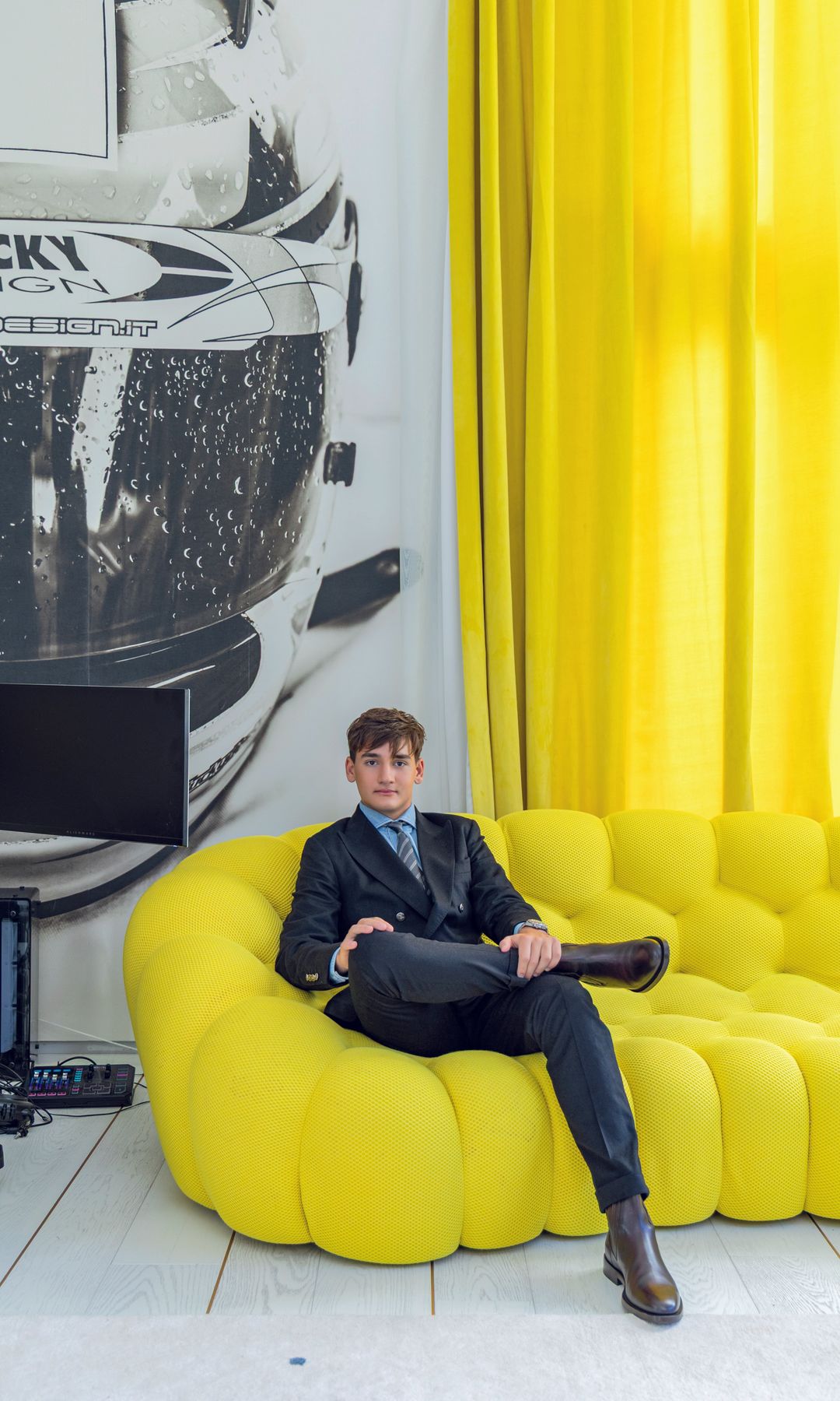 Hombre joven en traje y corbata, sentado en sofá amarillo en una sala con cortinas amarillas.