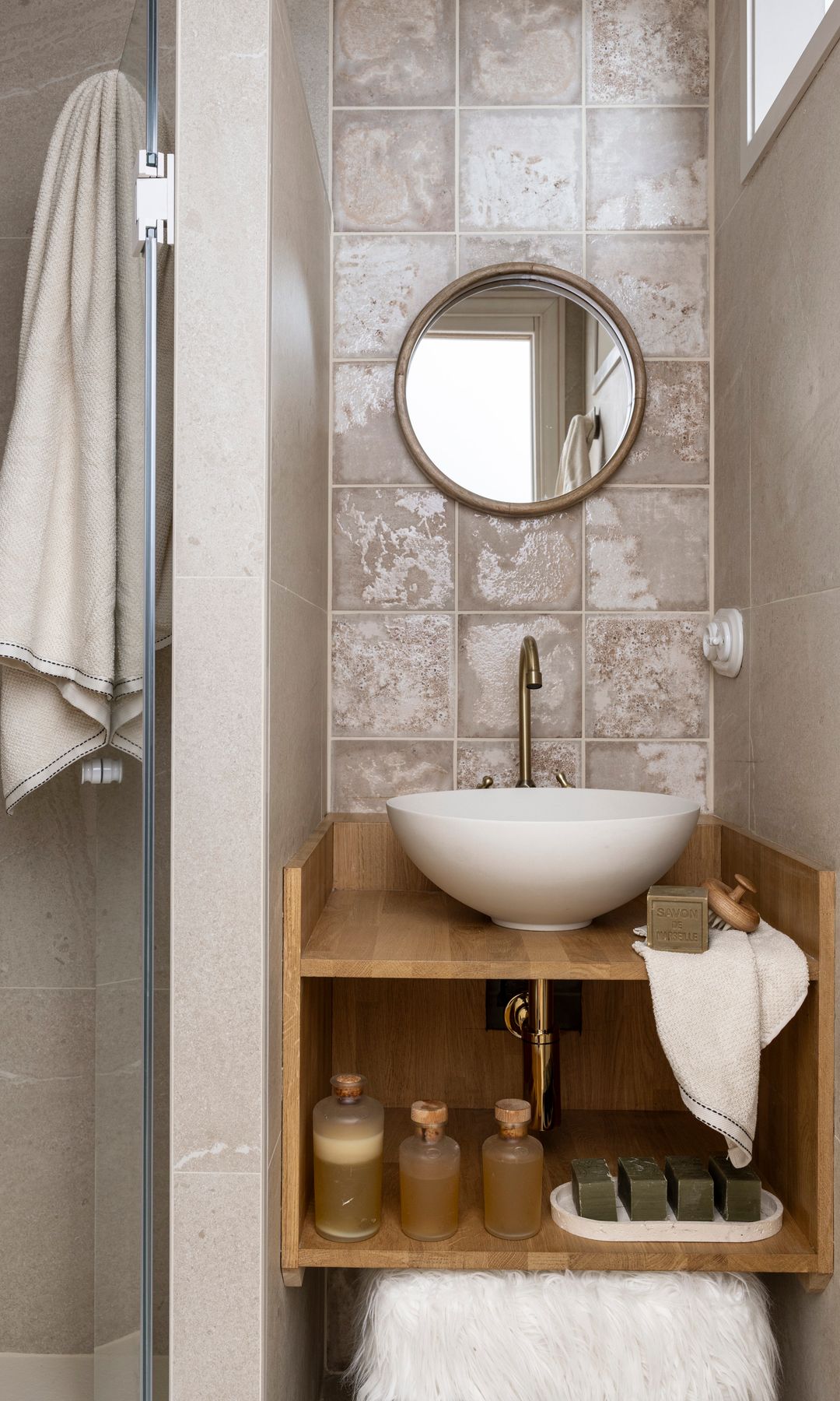 Baño pequeño con pared de azulejos beiges brillantes, espejo redondo, lavabo redondo sobre la encimera de madera, mueble de madera abierto con jabones, toallas y productos de higiene