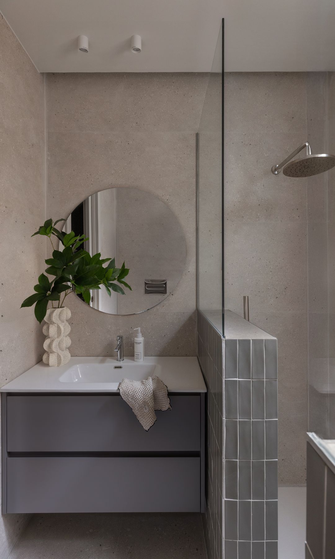 Baño pequeño, con ducha con medio murete de azulejos y cristal separando la ducha del lavabo, mueble volado gris, espejo redondo, jarrón con ramas verdes