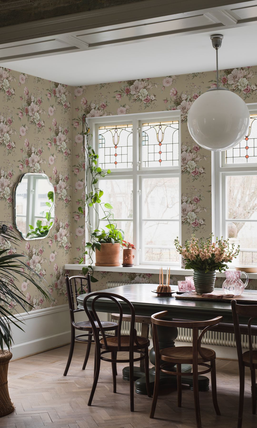 Comedor con papel pintado de flores, espejo, mesa de madera oscura, plantas en la ventana, sillas de madera antiguas