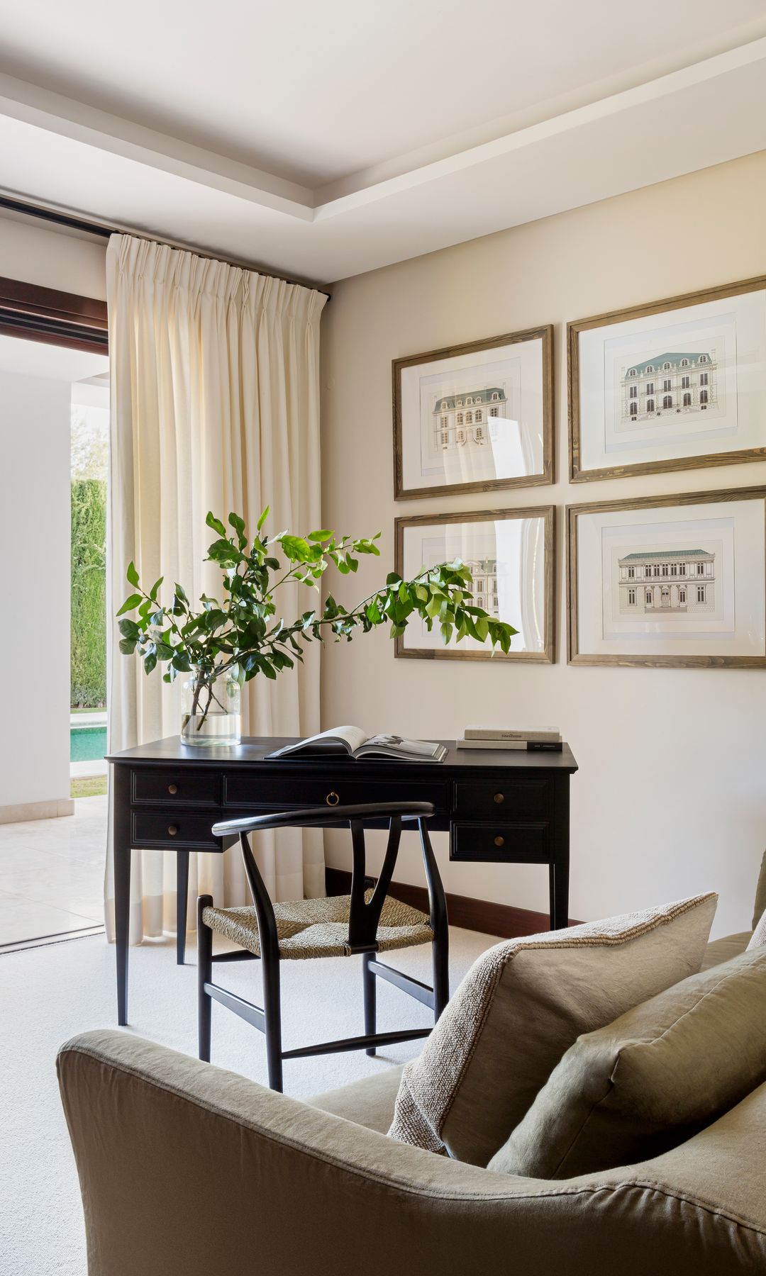 Despacho en casa junto a una salida al jardín, con escritorio negro, silla negra, cuadros en la pared, jarrón de cristal con planta y sofá cama 