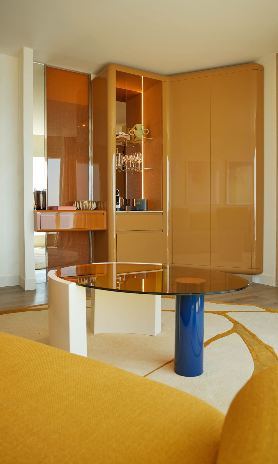 Salón con alfombra, mesa de centro de cristal con el pie azul, armario amarillo brillante, con zona abierta con baldas y trasera de cristal