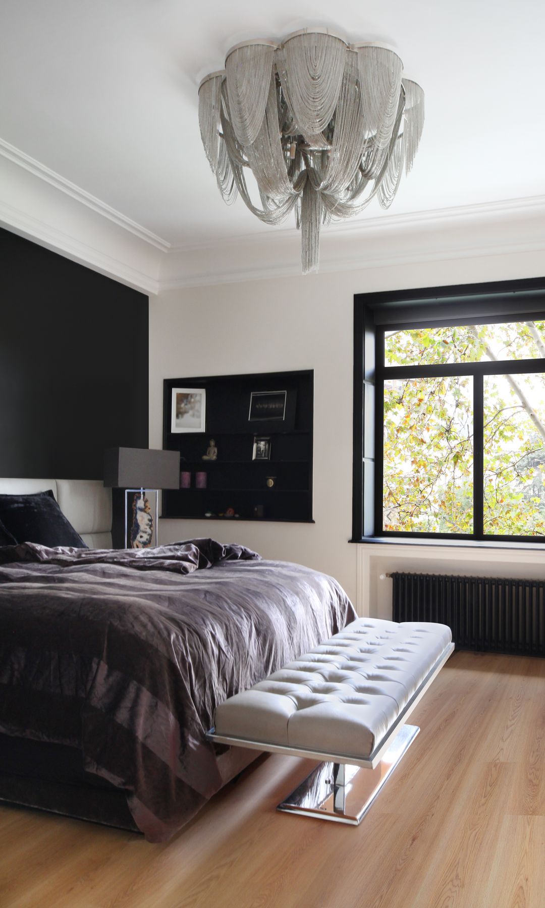 Dormitorio con gran ventanal negro, suelo de madera, rosa de cama gris oscuro, banqueta blanca a los pies de la cama, tablón negro en la pared, lámpara de techo 