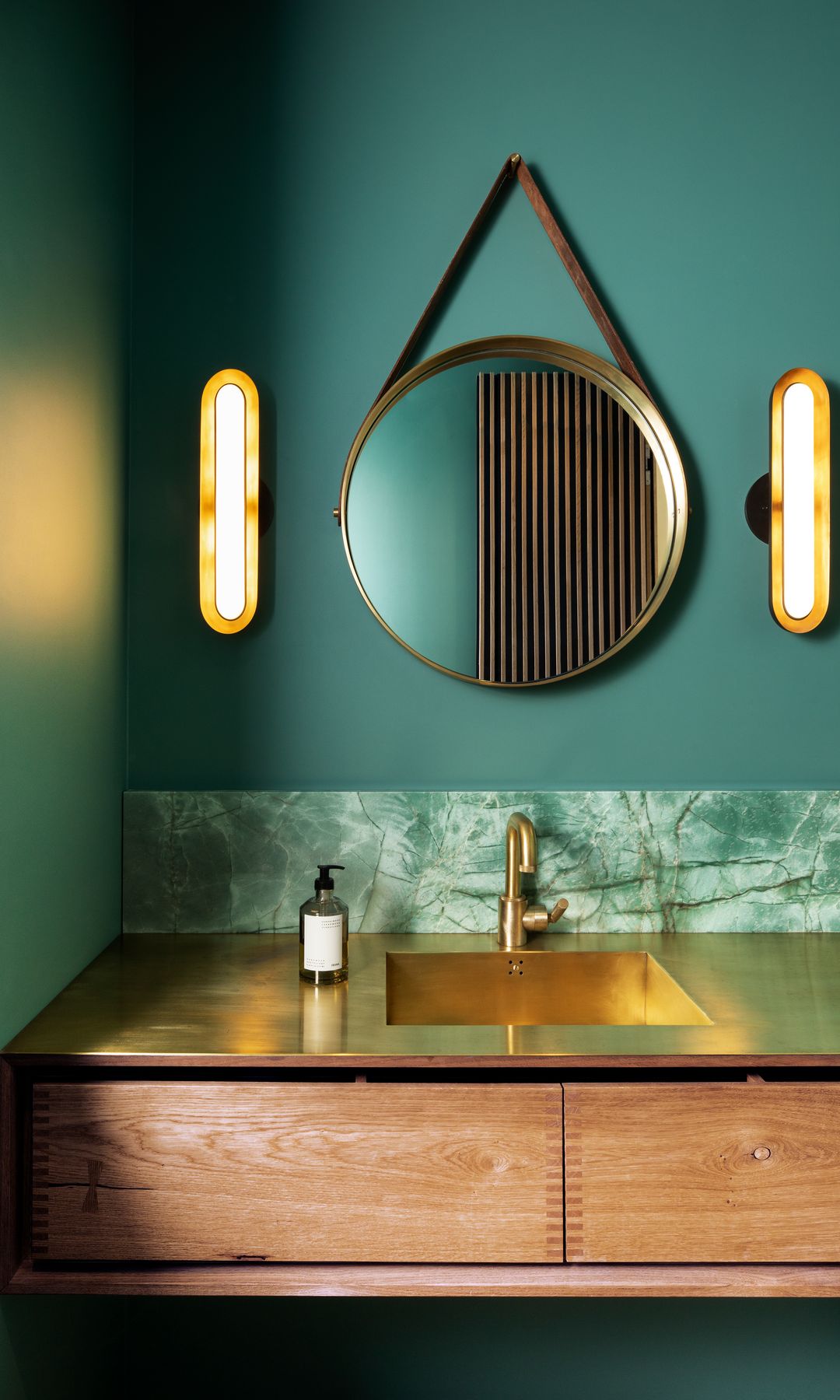 Baño con paredes verdes oscuras, zócalo verde, mueble de madera, encimera dorada, espejo redondo y apliques de pared