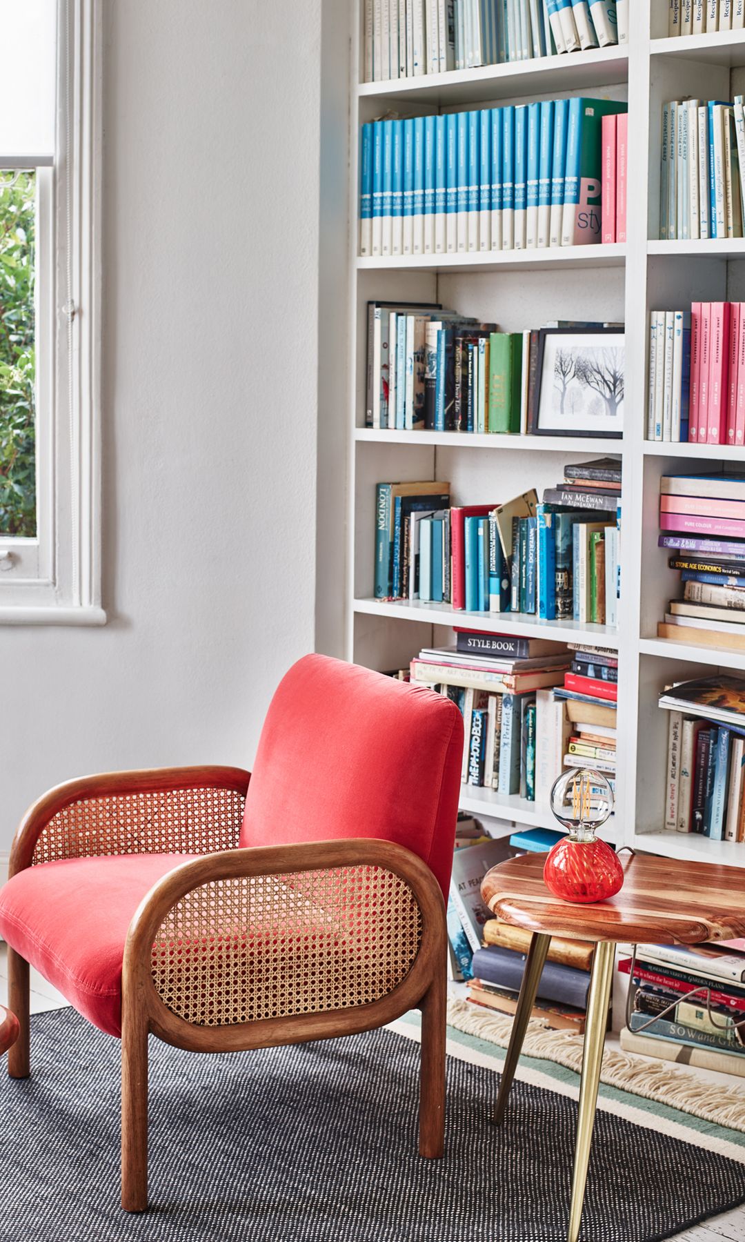 Rincón de lectura con librería blanca, butaca de rejilla, alfombra gris, ventana, mesas auxiliares con las patas doradas 