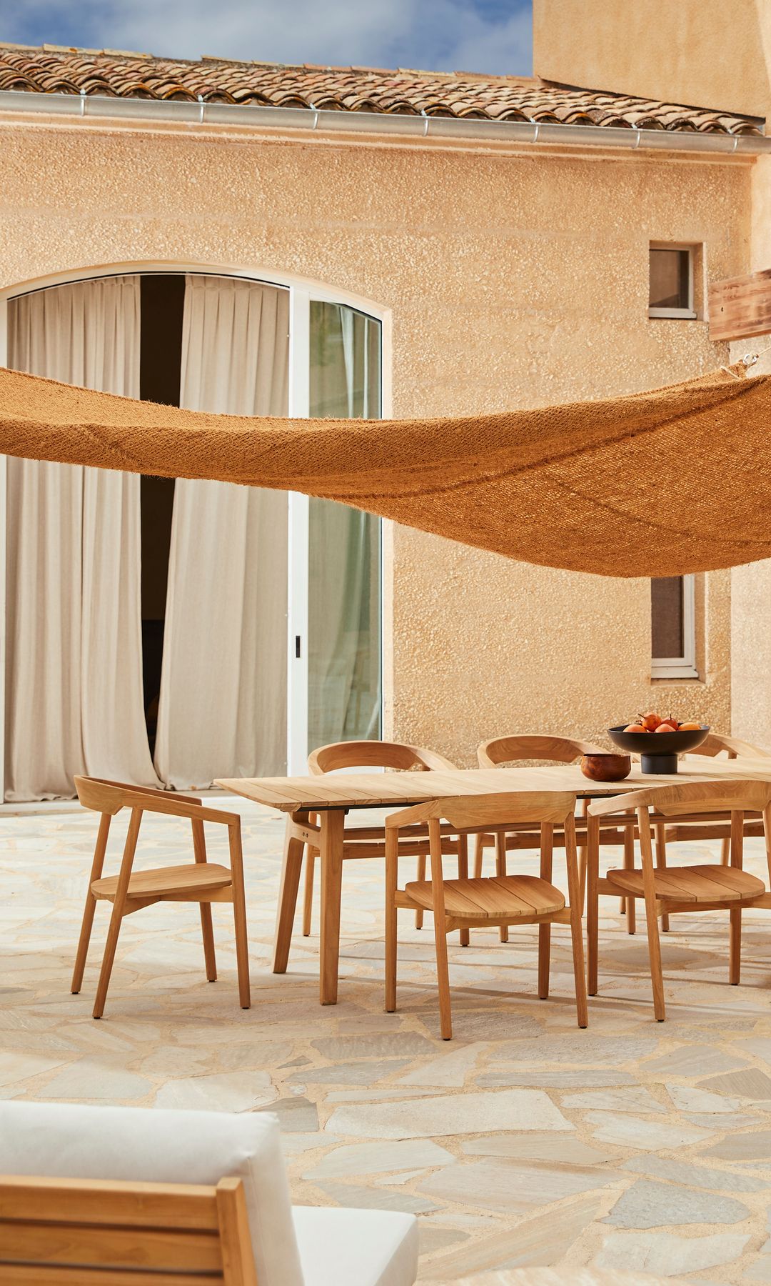 Comedor exterior con vela a modo de tildo, sillas de madera y suelo de piedra