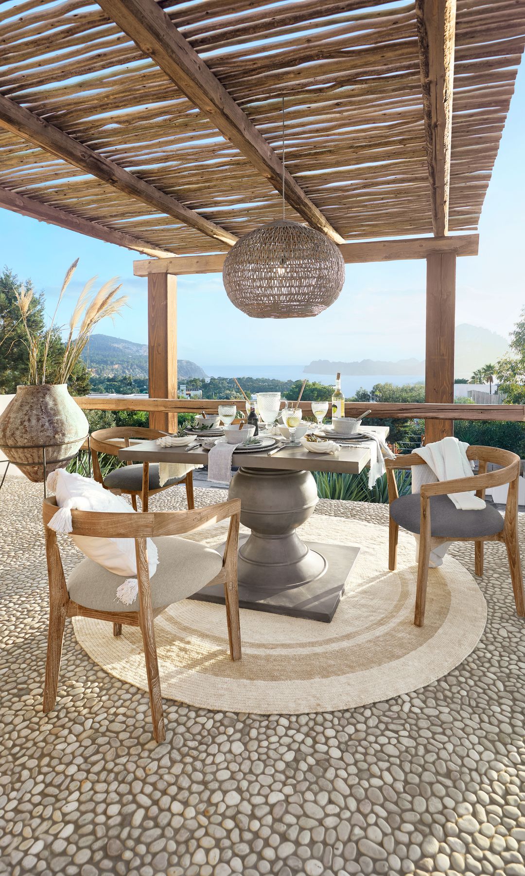 Comedor de verano con pérgola de caña, suelo de piedra, mesa con el pie gris, sillas tapizadas, lámpara de techo de fibras vegetales