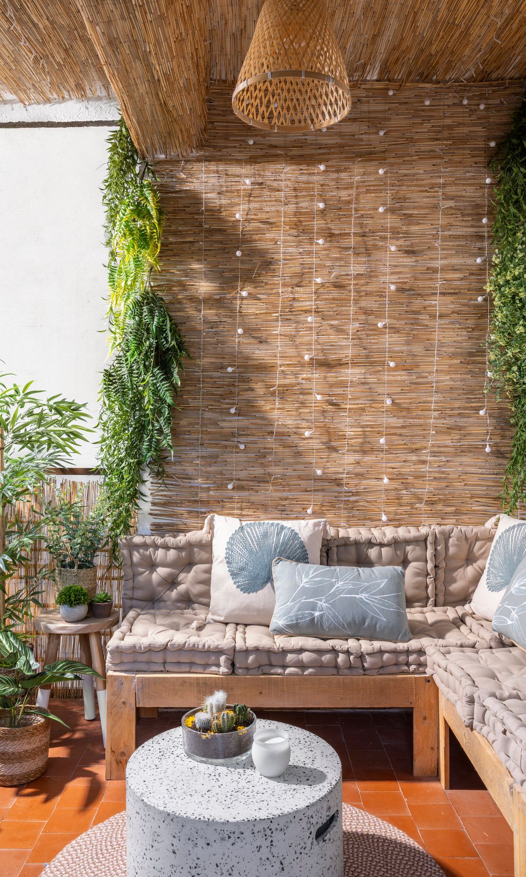Terraza con techo y paredes de paja, lámparas de fibras vegetales, sillones de madera con cojines, mesa de centro redonda, plantas colgantes y en el suelo 