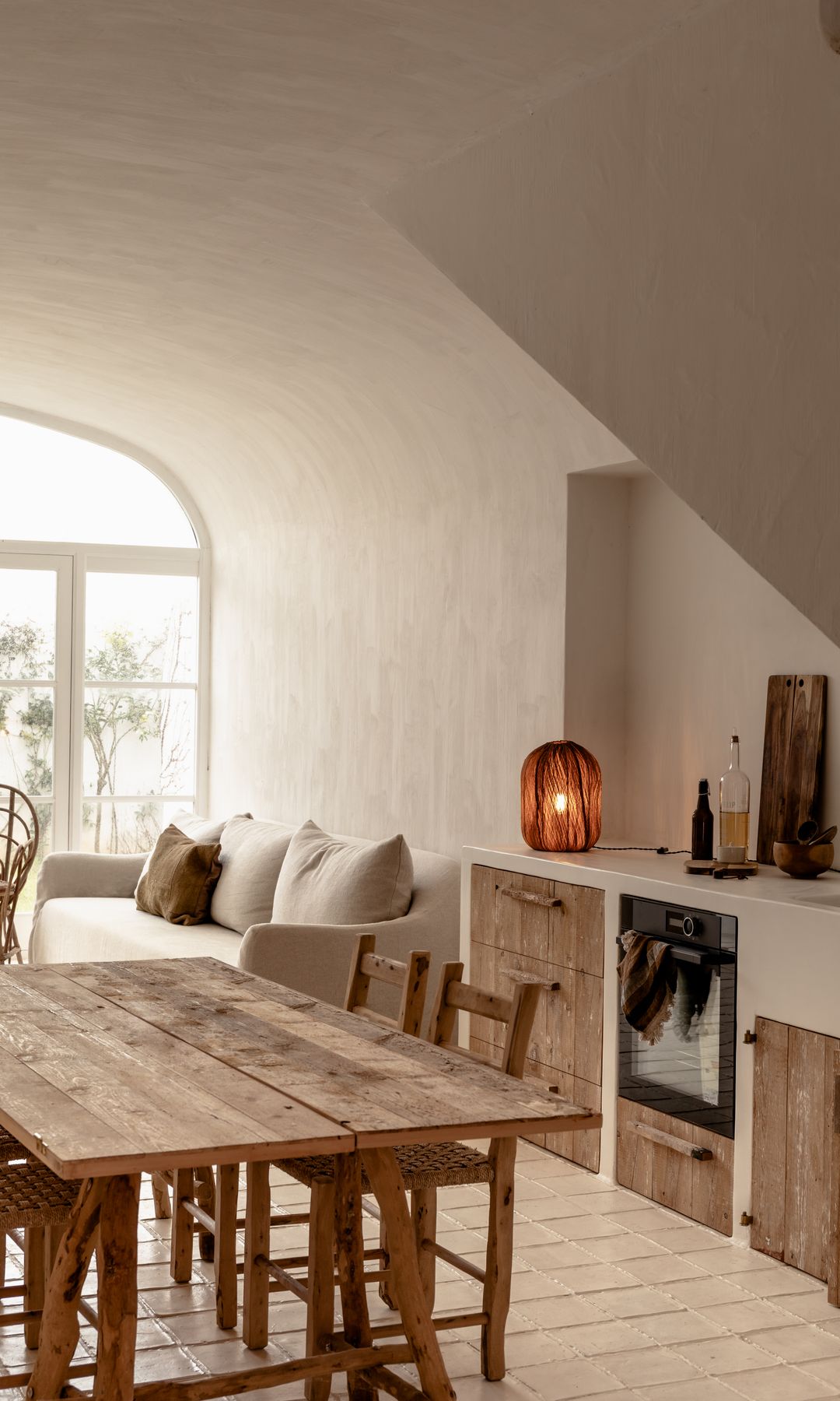 Cocina abierta al salón y al comedor, de estilo rústico con mesa de madera, sofá blanco, lámpara de sobremesa de fibras vegetales 