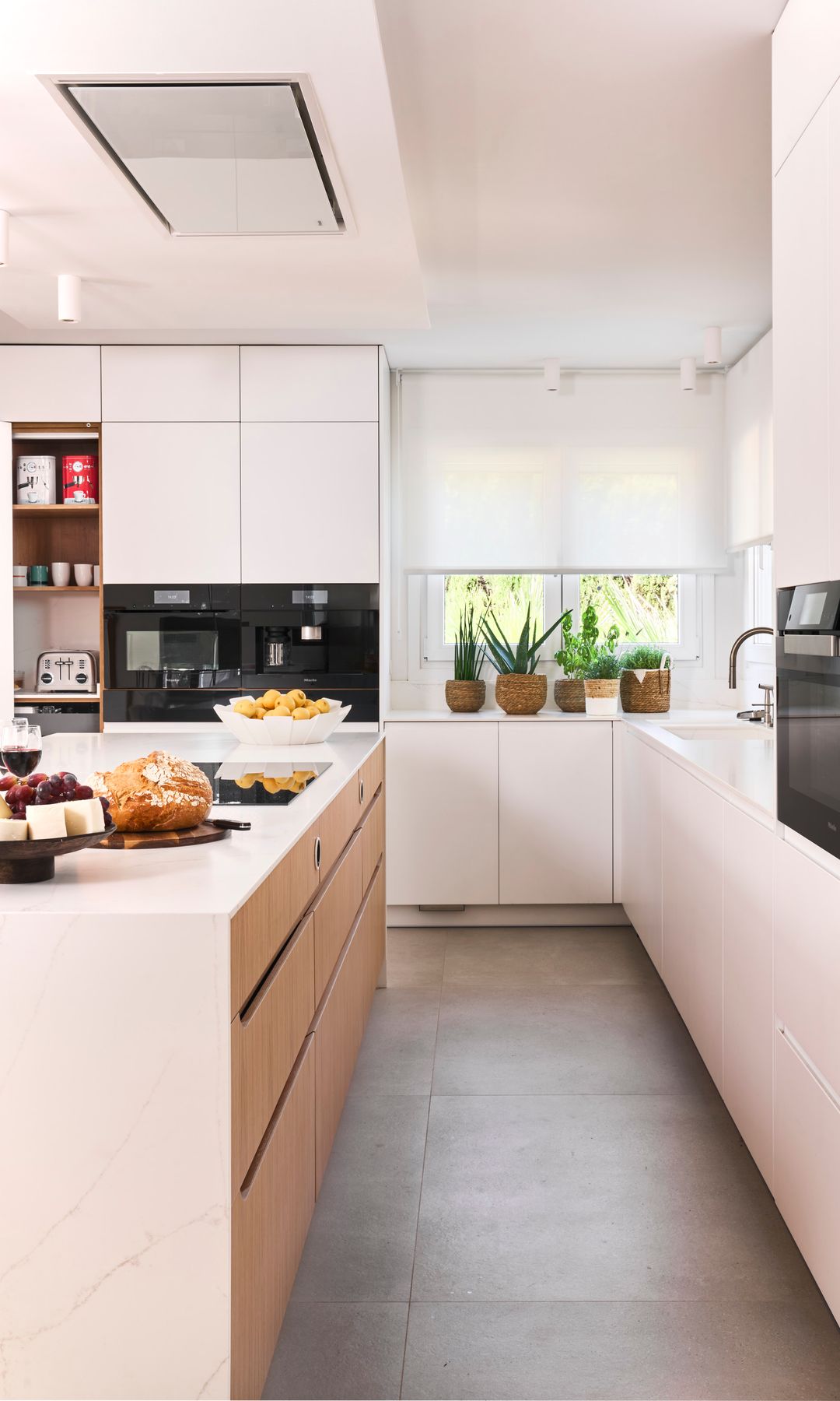 Cocina blanca con módulos hasta el techo, isla de cocina, campana en el techo, estor en la ventana, electrodomésticos negros, suelo gris