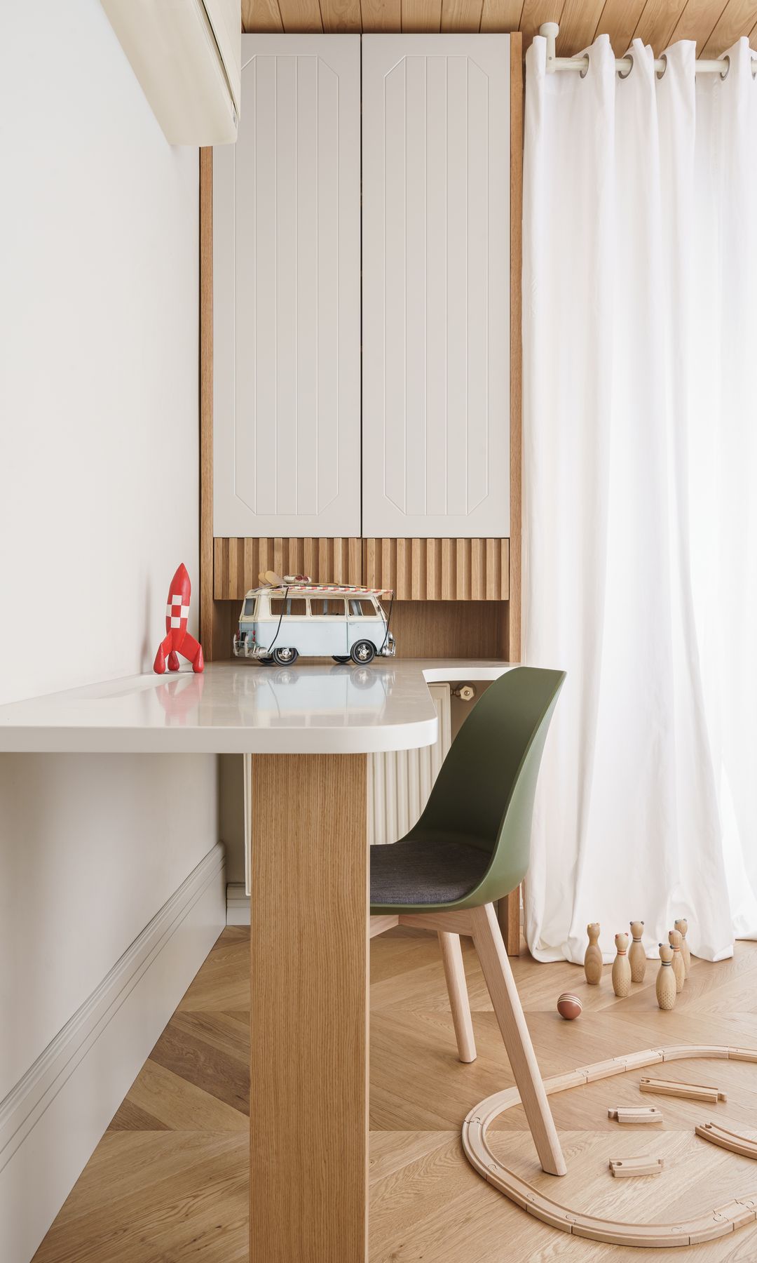 Habitación infantil con mesa de escritorio blanca con pie de madera, armario, cortinas blancas, radiador bajo el mueble, juego infantil de madera en el suelo 