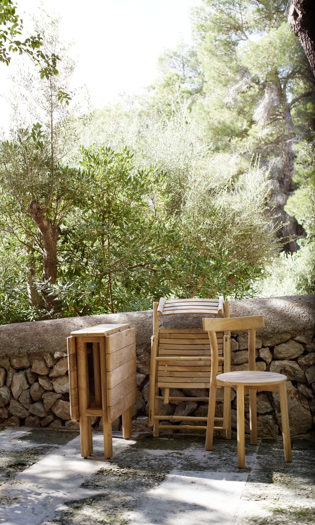 Mesa plegable de madera con taburete y sillas apoyados en un muro en un jardín 