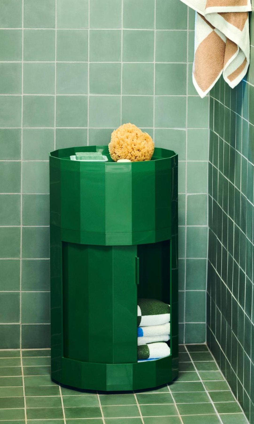 Baño con azulejos verdes, contenedor verde de plástico con repisa en la parte superior y espacio interior para guardar las toallas