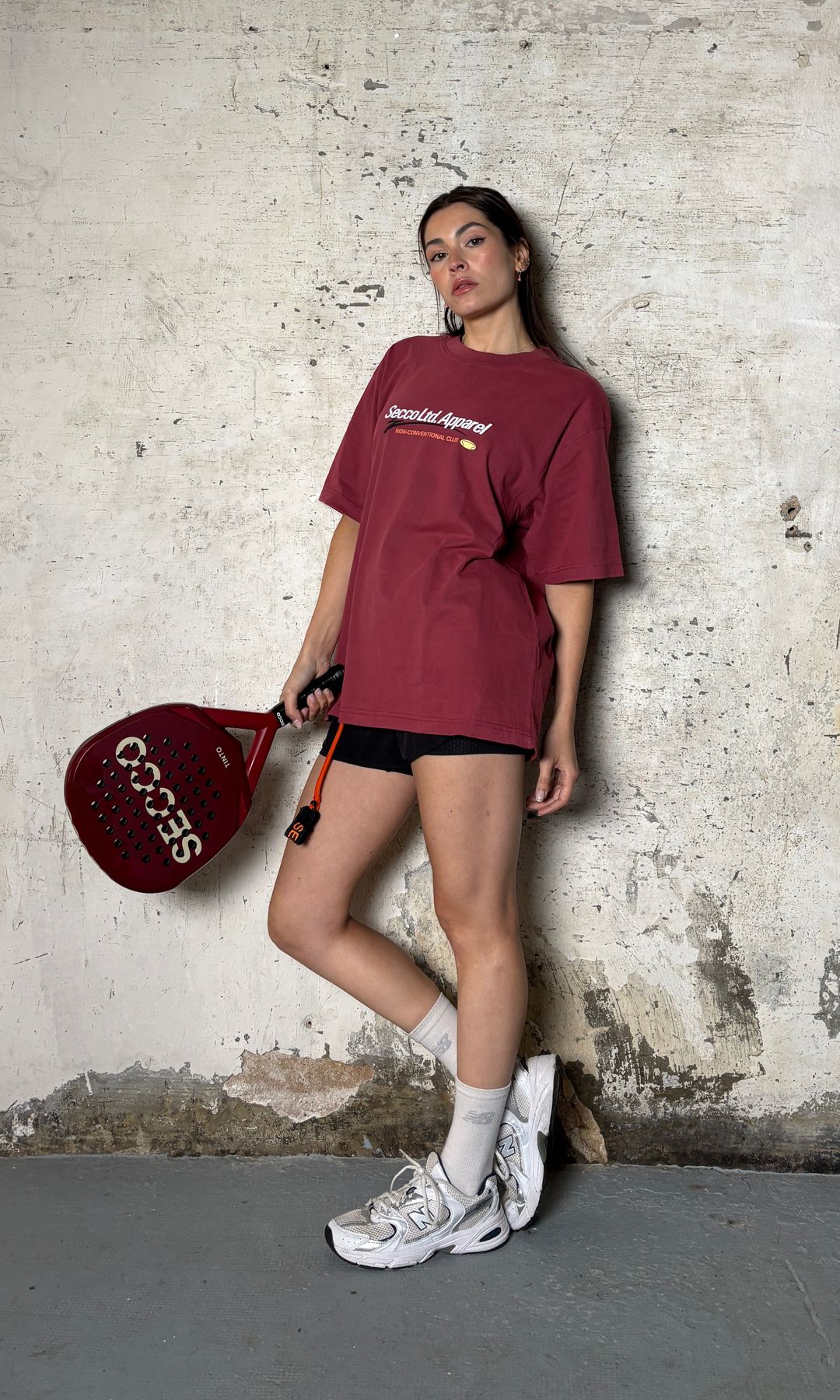 Mujer con raqueta de pádel, camiseta roja y zapatillas blancas posando contra pared deteriorada.