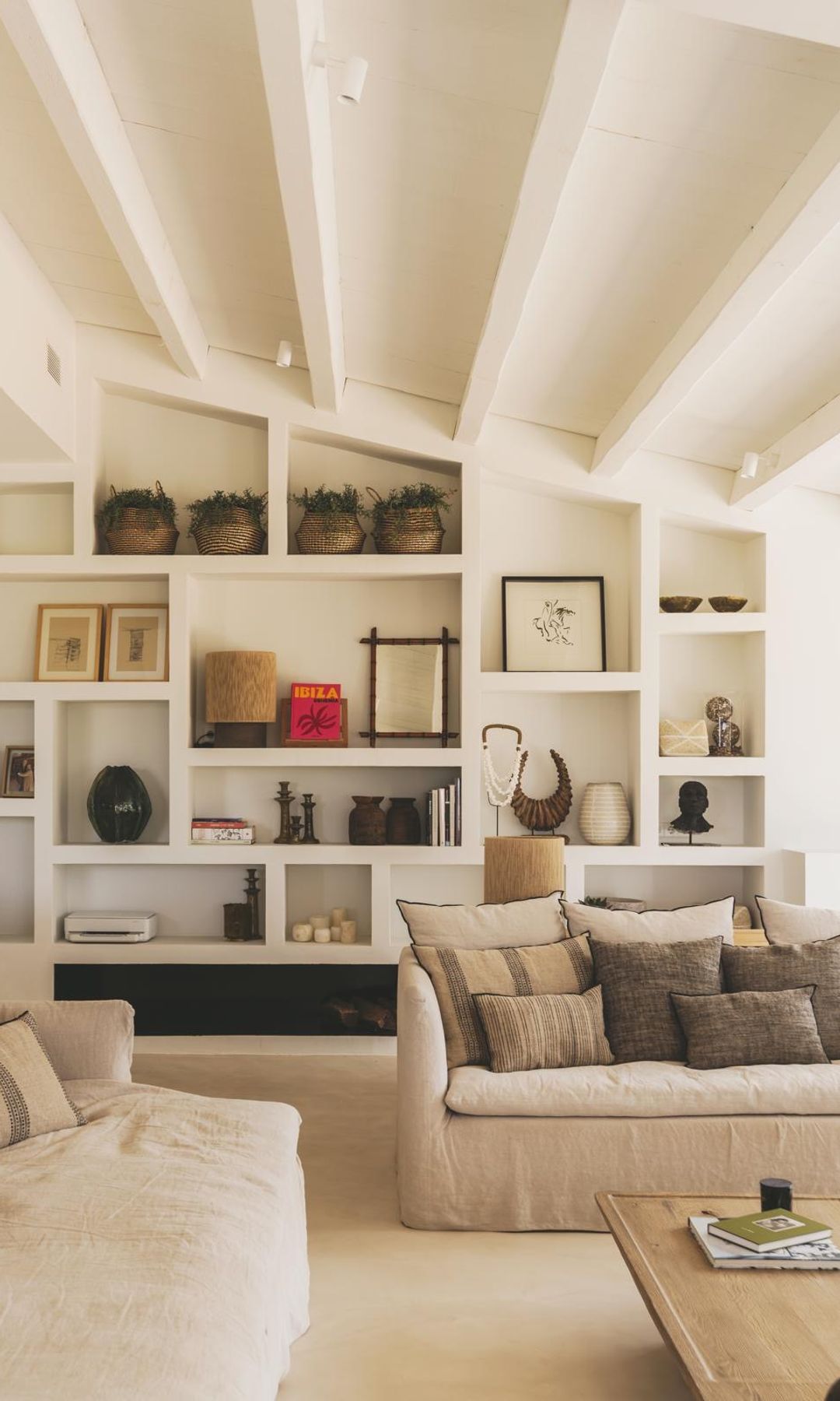 Salón con estantería de obra, sofás con fundas de lino beige, mesa de centro de madera clara, cojines en el sofá, vigas en el techo 