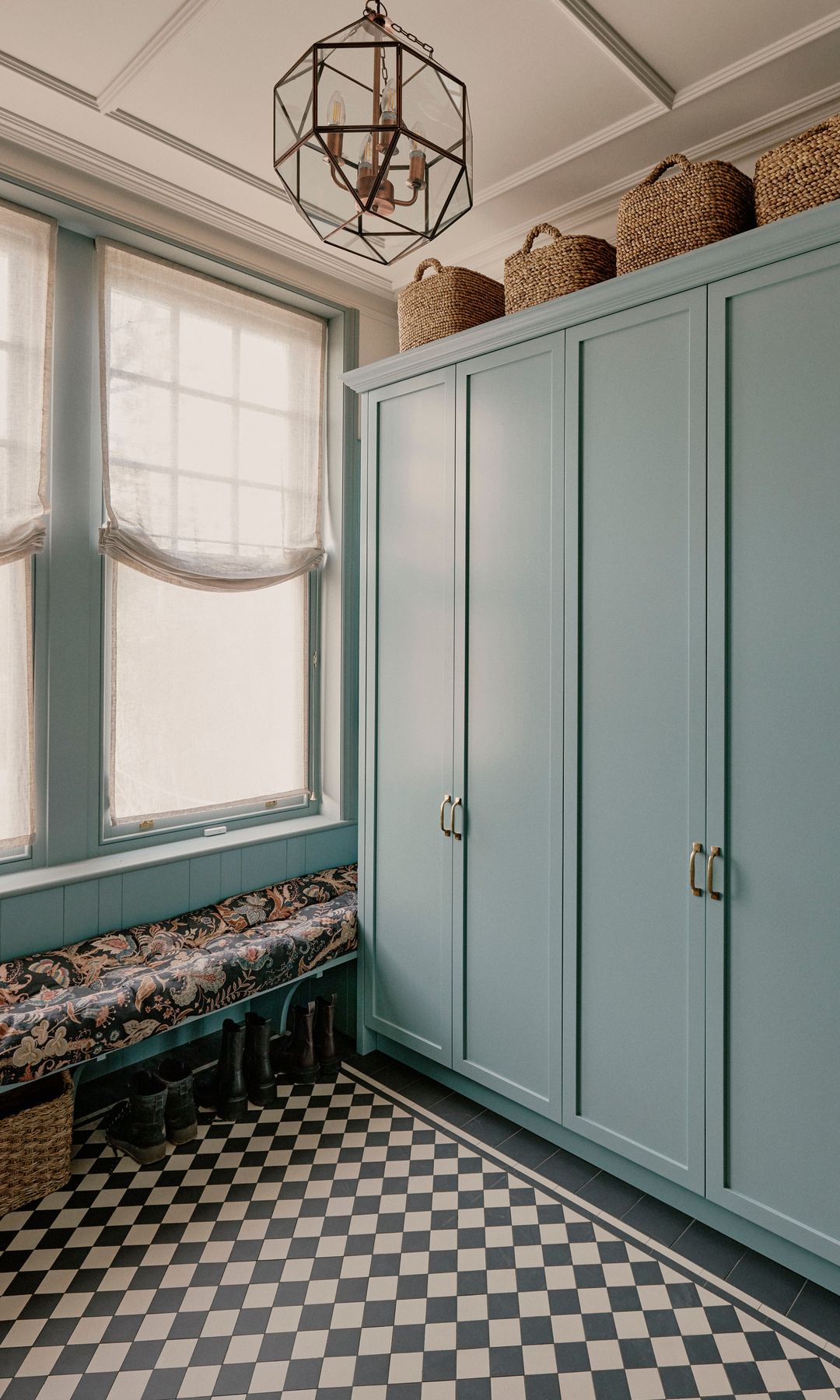 Lavadero con fregadero, suelo de damero blanco y negro, armario azul claro, banco con tela de flores negras, estores blancos recogidos, cestos en lo alto del armario