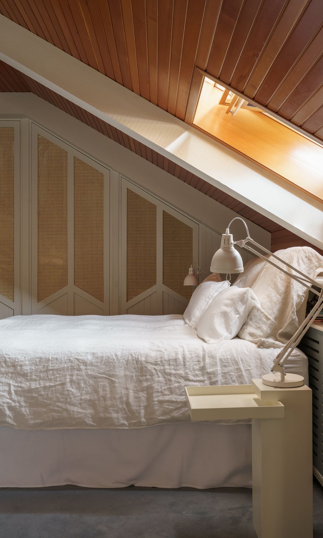 Dormitorio abuhardillado con techo de madera, ventana de techo y armario en desnivel con puertas blancas y de rejilla