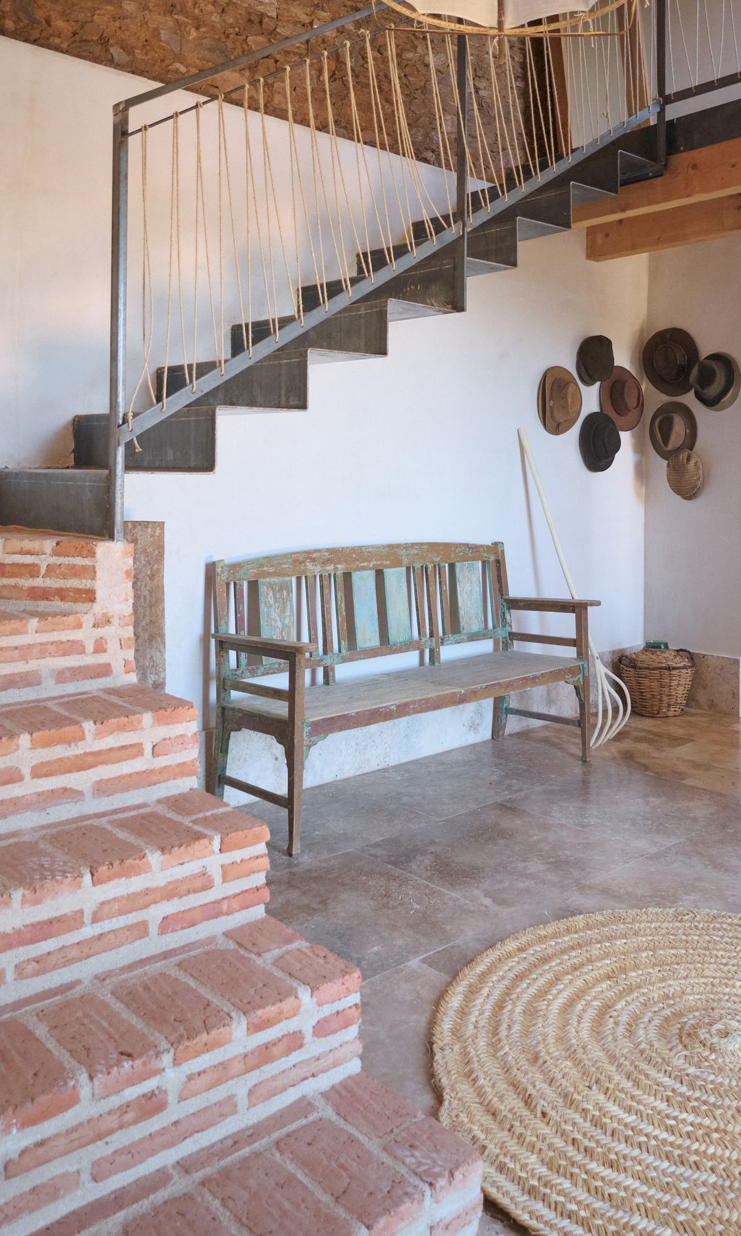 Recibidor de una casa de campo con alfombra redonda de fibras naturales, escalones de ladrillo, banco, sombreros colgados de la pared, cesto en el suelo