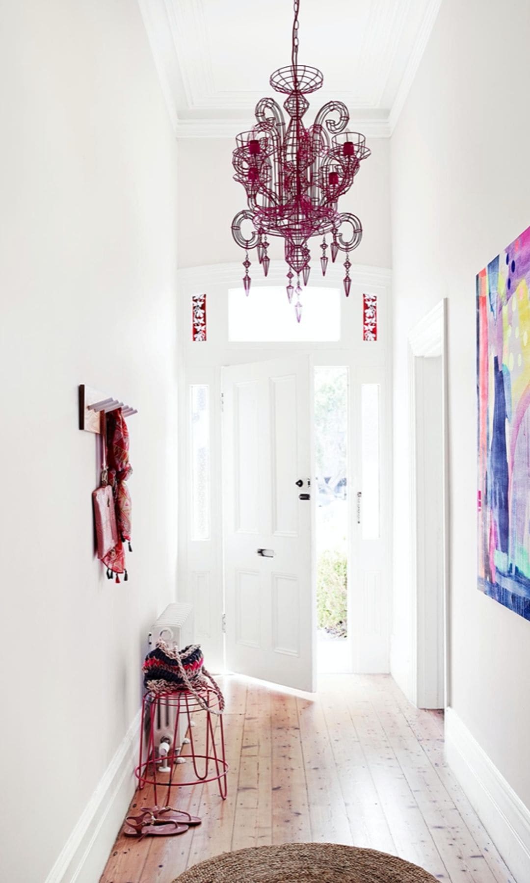 Pasillo que da a la entrada de la casa, con paredes blancas, cuadro de colores colgado en la pared, suelo de madera, alfombra, lámpara de techo roja, perchero de pared rojo y taburete metálico rojo