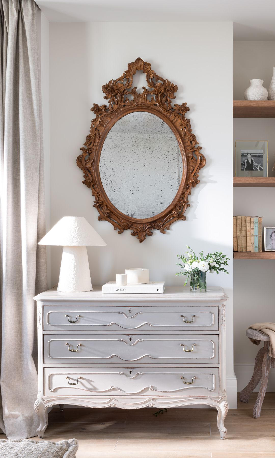 Rincón con cómoda antigua, espejo con el marco dorado, lámpara de sobremesa, hornacina con baldas de madera en la pared con fotos