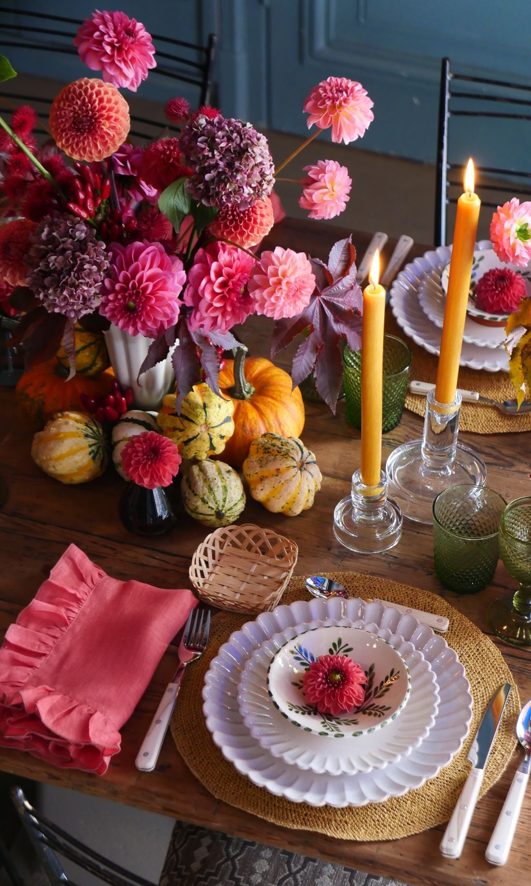 Mesa de comedor vestida con arreglo floral en tonos rosas, platos blancos con bordes labrados, salvamanteles, servilleta rosa, velas, candelabros, calabazas