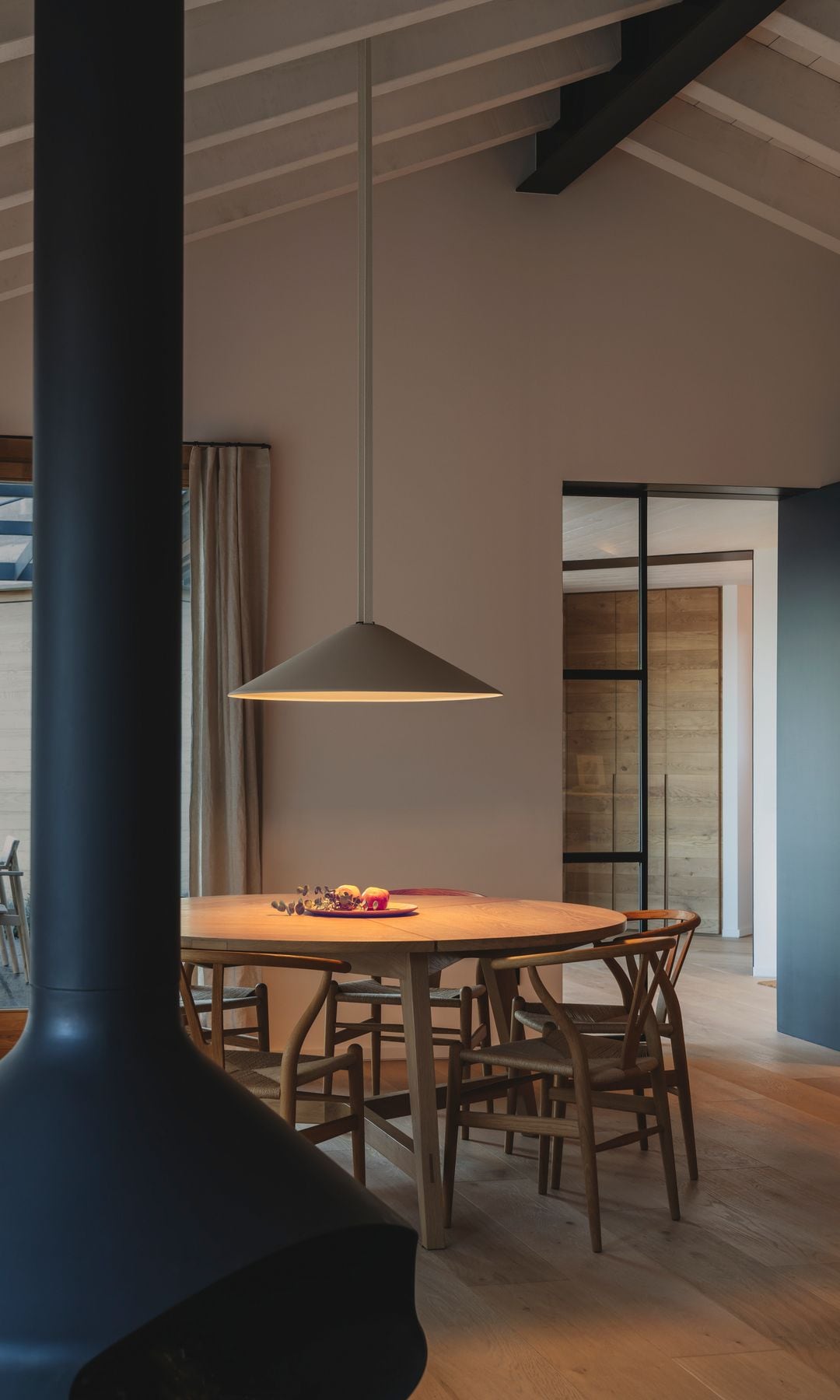 Comedor con chimenea y lámpara de techo encendida, mesa de comedor redonda de madera, sillas de madera, vigas en el techo