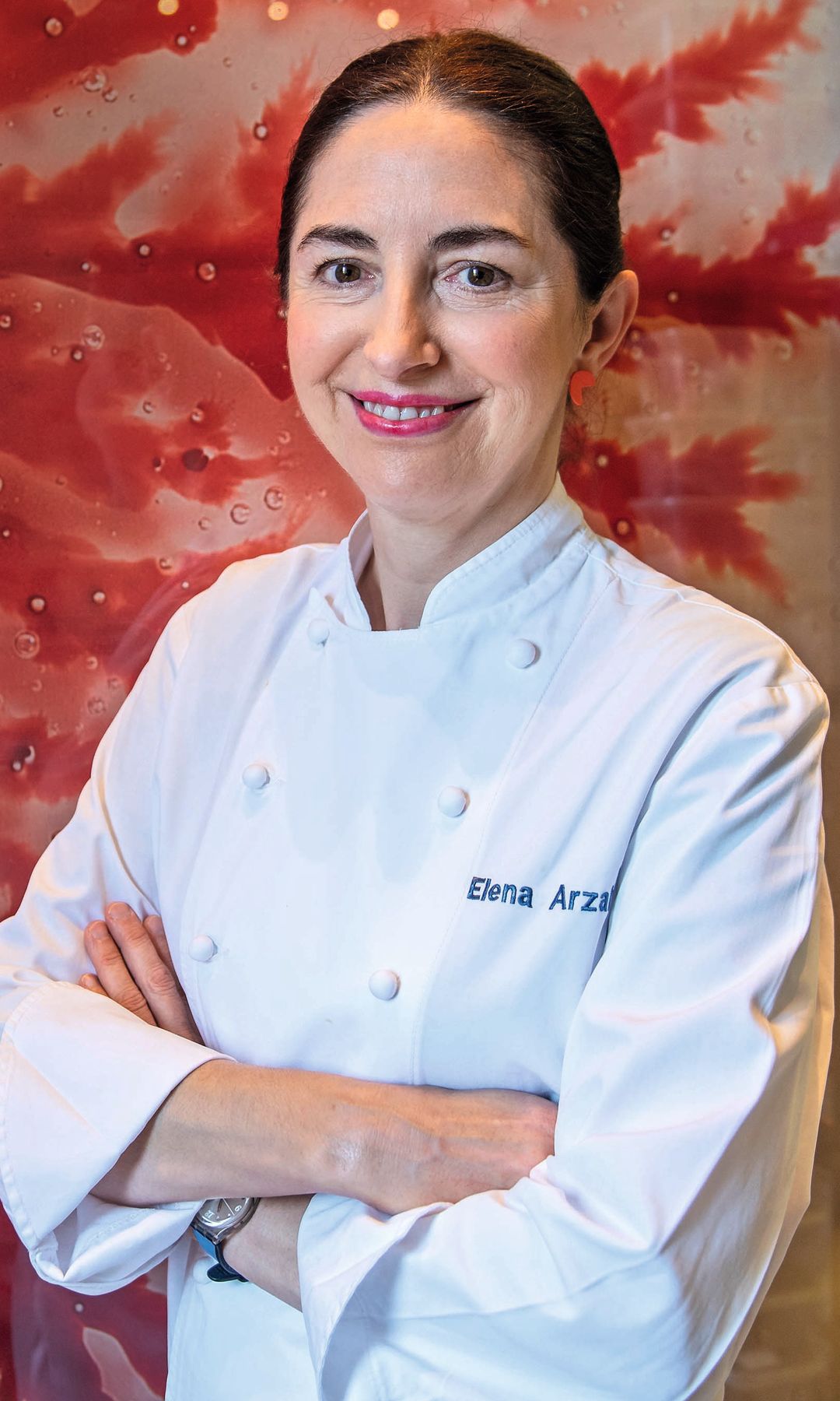 Elena Arzak, con uniforme de chef blanco, sonríe con brazos cruzados frente a un mural rojo.