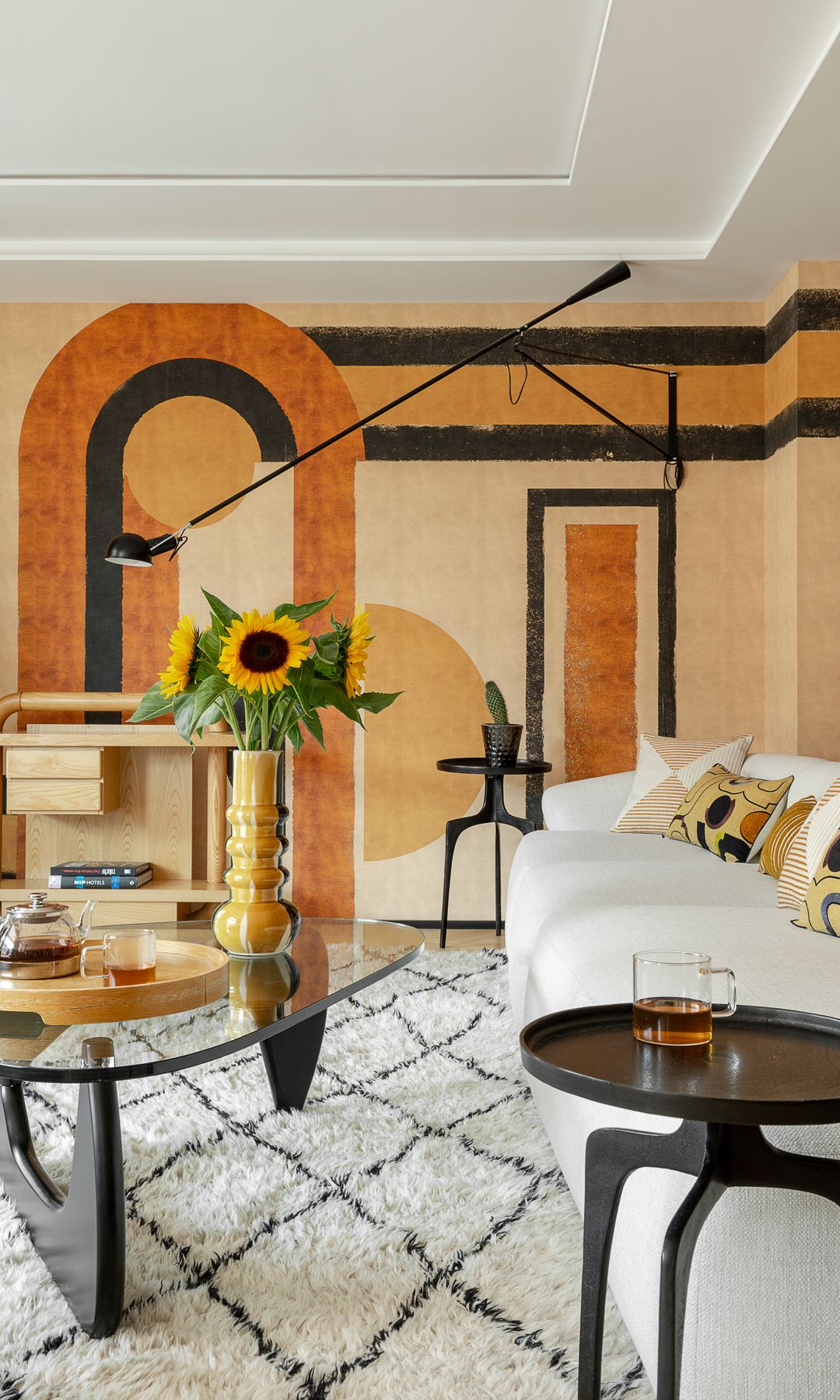 Salón con sofá blanco, mural de papel pintado en la pared, mesa de centro de cristal, mesa auxiliar negra, alfombra, jarrón con girasoles, techo con molduras