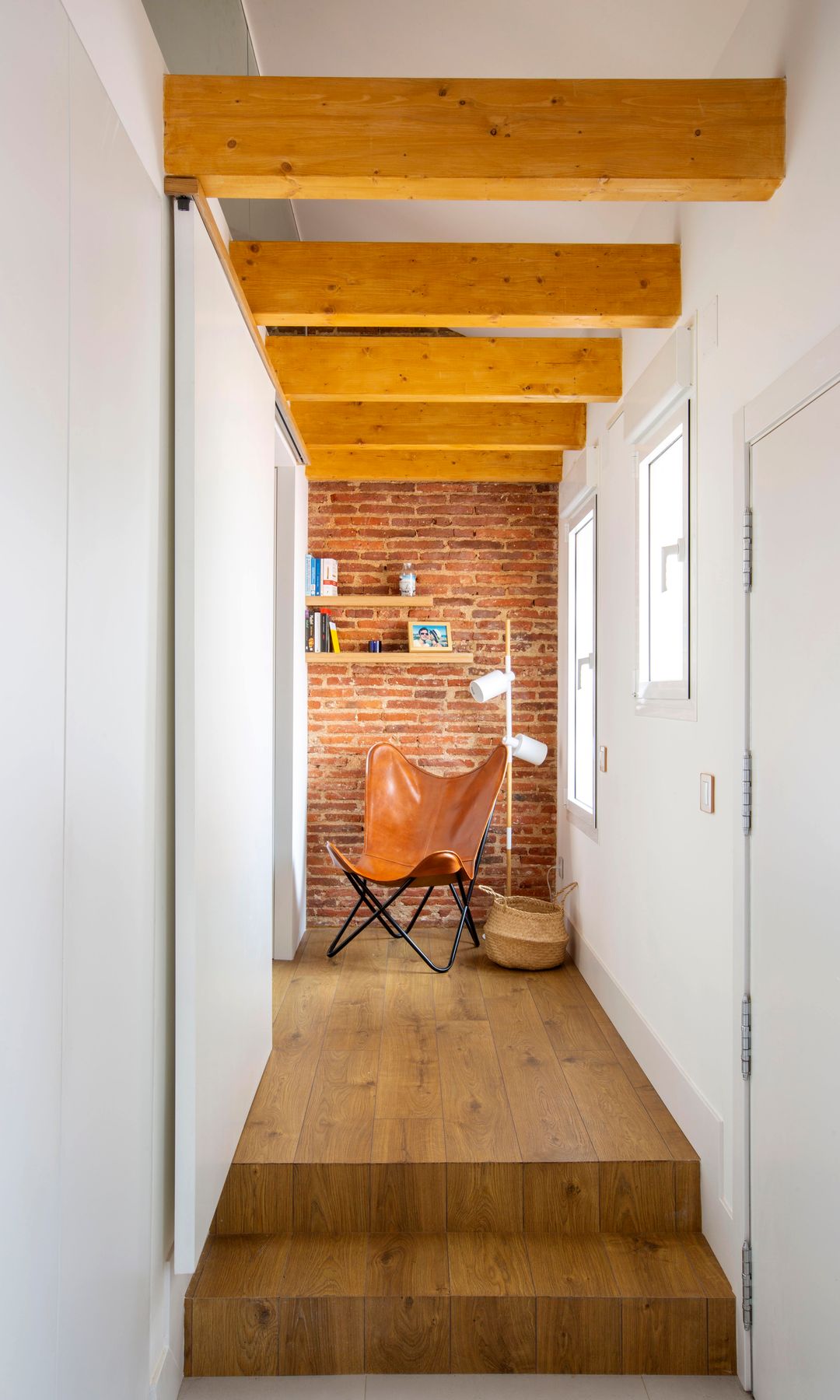 Pasillo con ventanas, suelo de madera, vigas de madera y rincón con butaca de cuero, pared de ladrillo y baldas