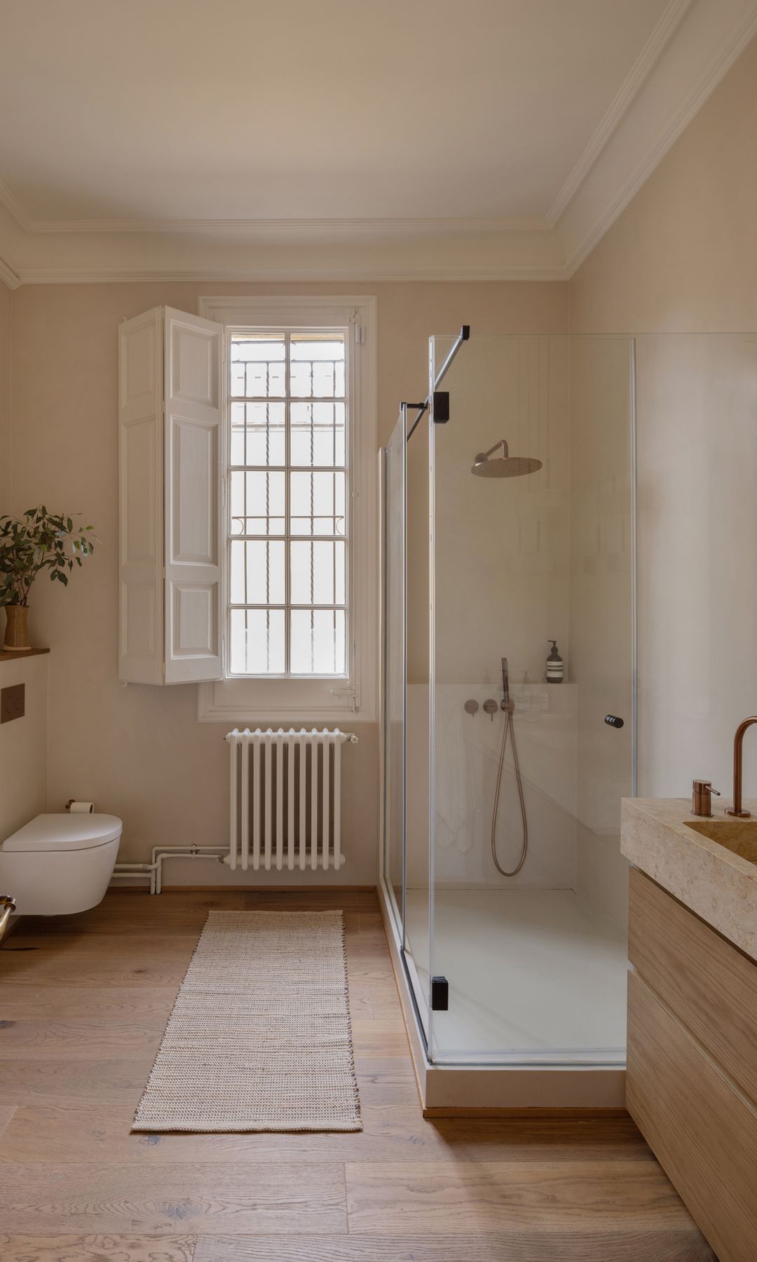 Baño con ducha XL con cabina de cristal, suelo de madera, alfombra, ventana, sanitario suspendido, balda murete sanitario con jarrón con ramas verdes