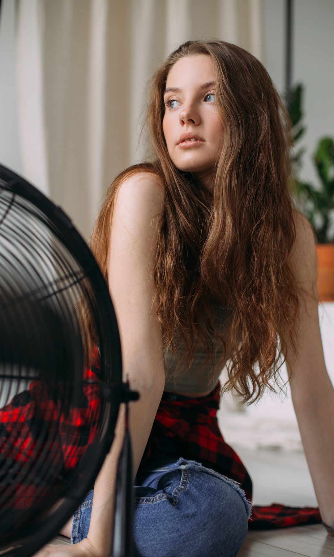 Chica joven en el suelo de su habitación con un ventilador negro delante.