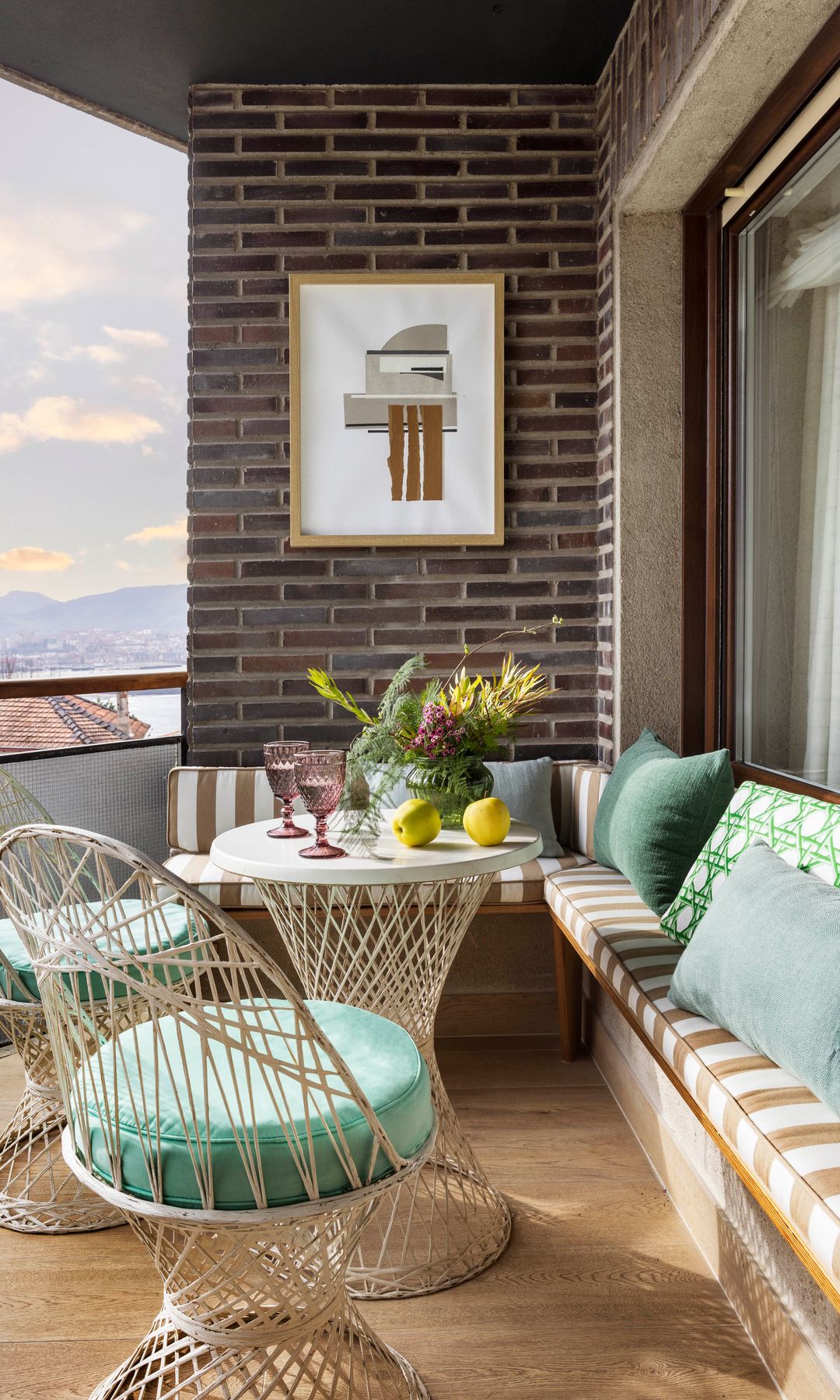 Terraza con banco con cojines asiento de rayas, mesa redonda de fibras vegetales, sillas a juego con asiento verde, cojines vedes sobre el sofá, cuadro en la pared