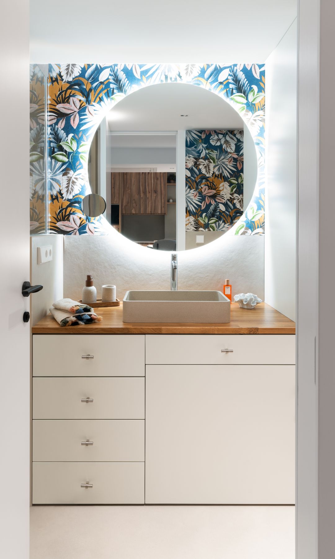 Baño con espejo redondo retroiluminado, papel pintado tropical, mueble blanco, encimera de madera