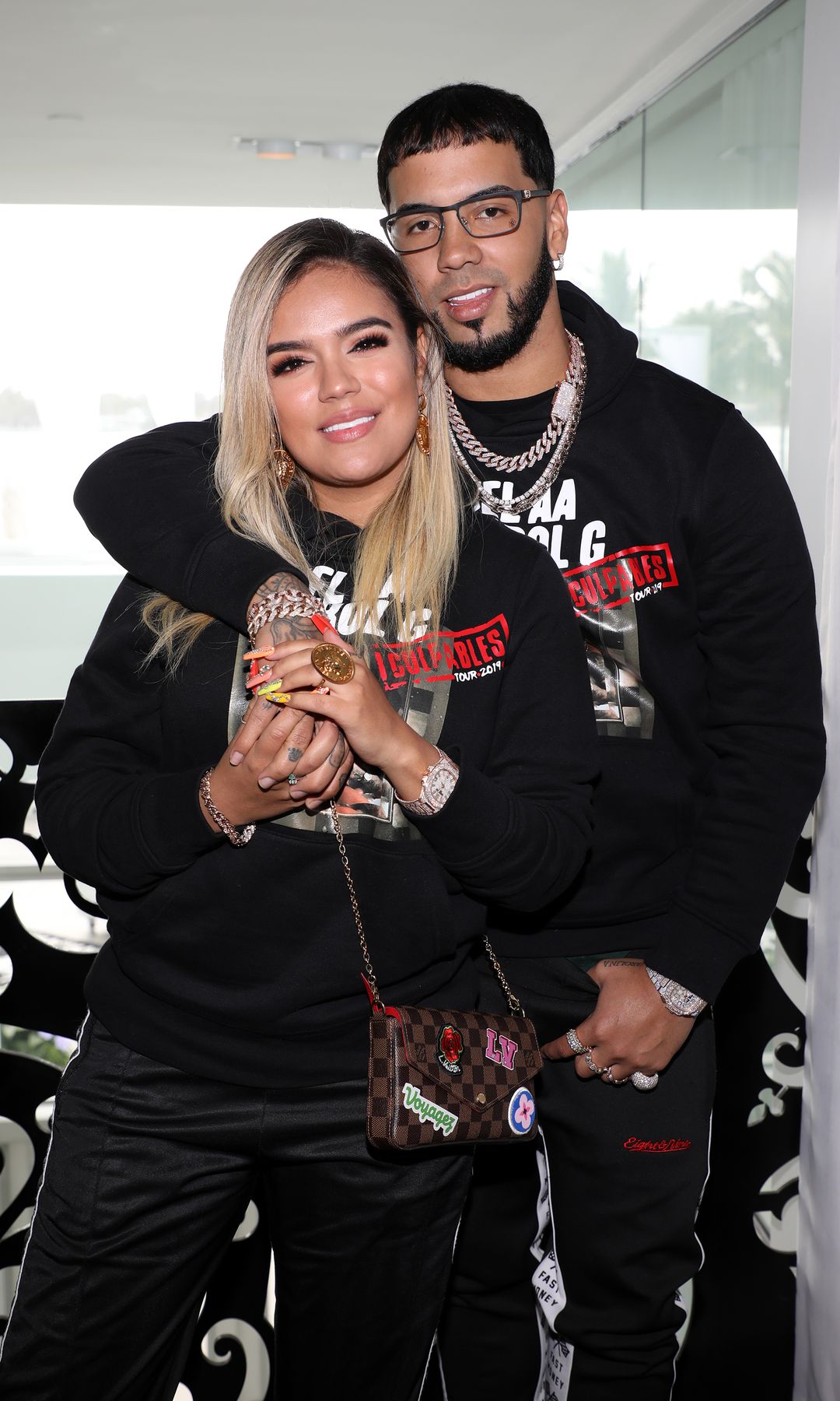 Karol G y Anuel AA ambos con ropa negra y joyas